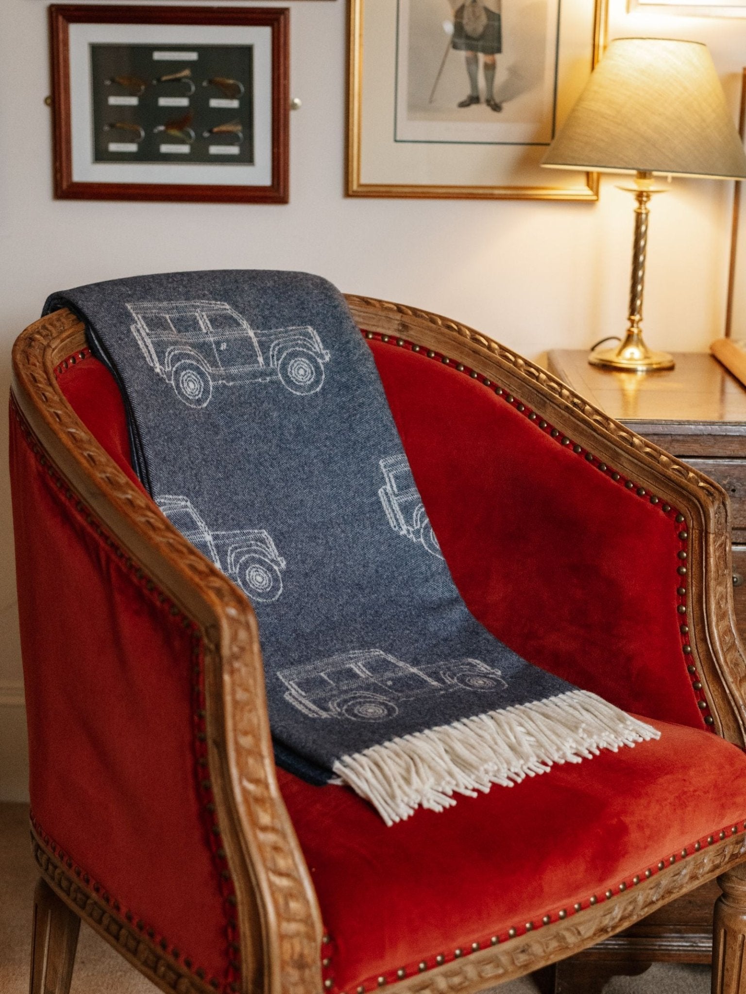 A red velvet armchair with wooden trim holds the Land Rover Defender Jacquard Throw by Campbell's of Beauly, a blue British wool blanket with white car motifs and fringe. Behind is a wooden desk, lit lamp, and framed art on a cream wall.
