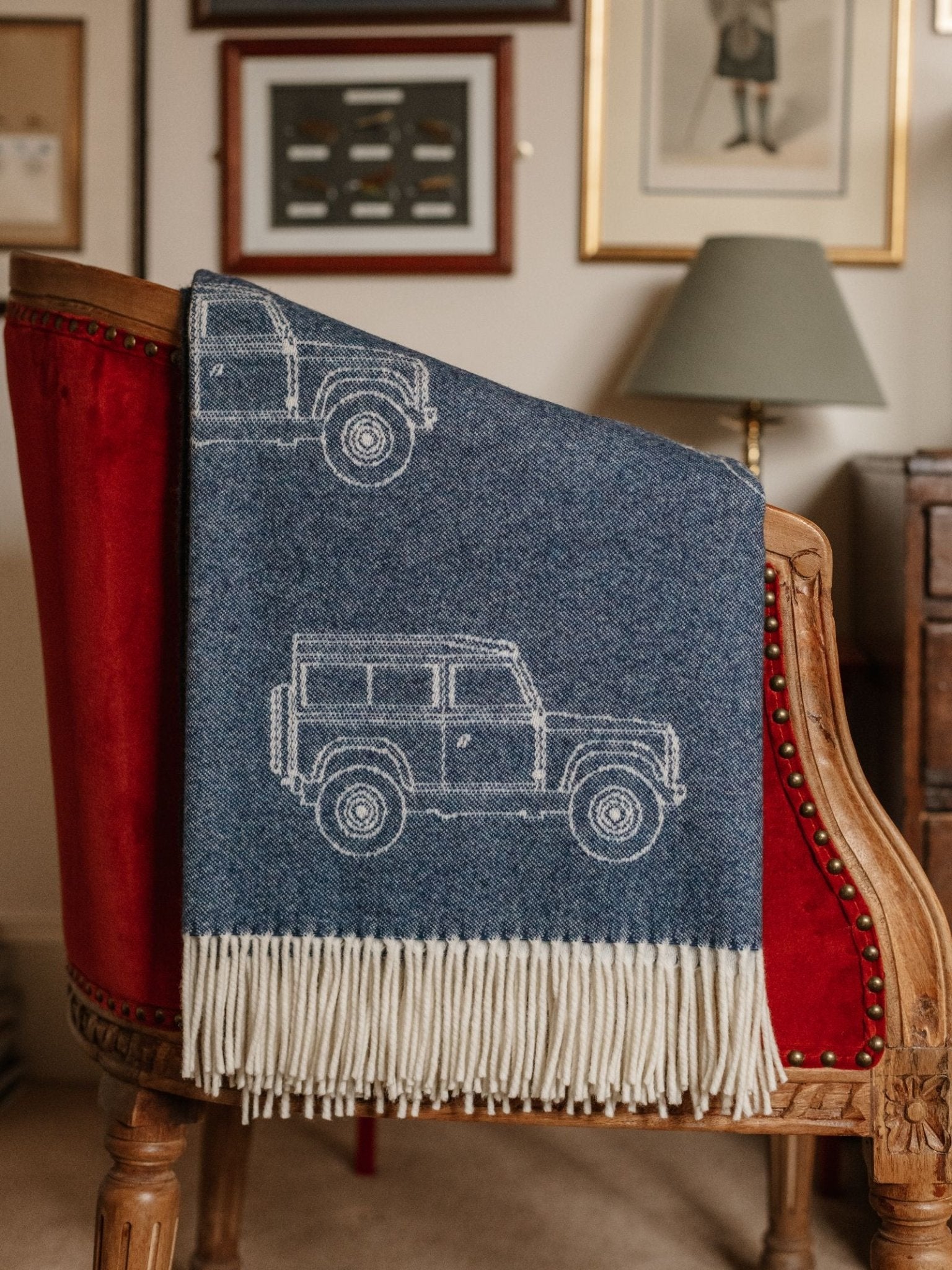 The Campbell's of Beauly Land Rover Defender Jacquard Throw, blue wool with white fringe and vehicle outlines, is draped over a red velvet wooden chair. Framed art and a table lamp appear in the background.