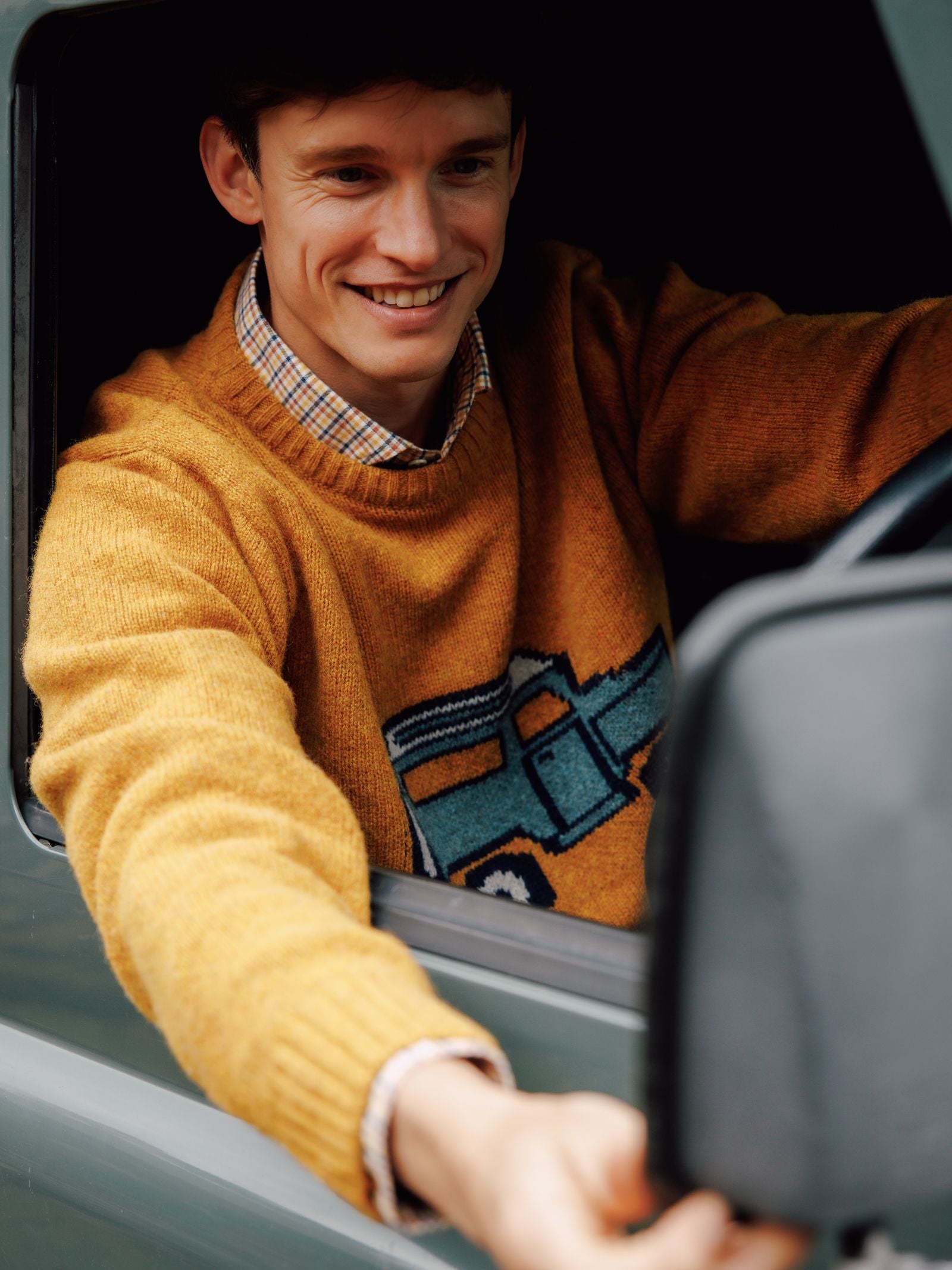 A smiling person in the Campbell's of Beauly Unisex Land Rover Defender Jumper adjusts a side mirror from a car window, showcasing classic knitwear with automotive flair.