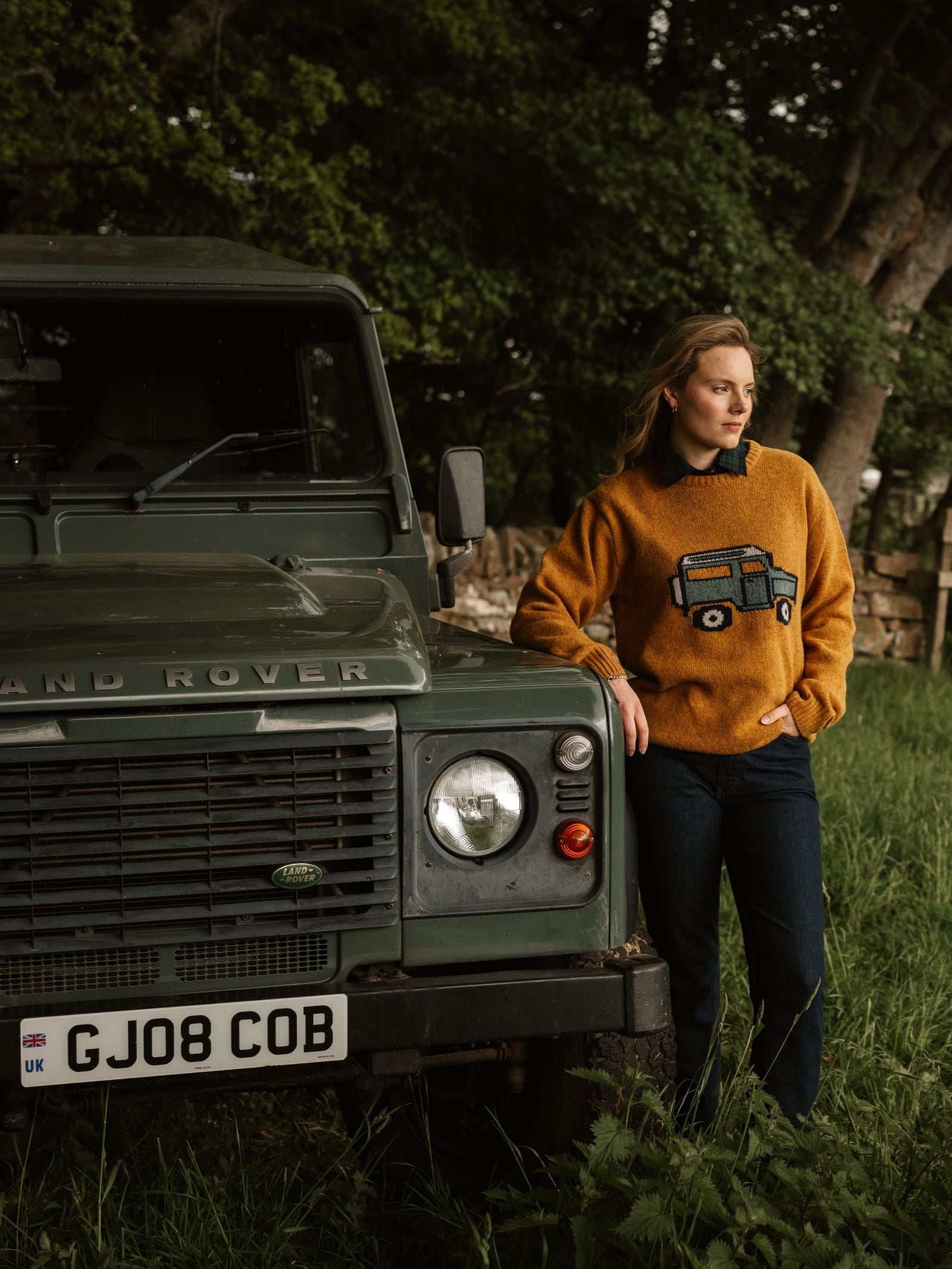 A woman in a mustard Unisex Land Rover Defender Jumper by Campbell's of Beauly stands next to a green Land Rover, set on grass with trees and a stone wall behind her—blending classic knitwear style with adventure.