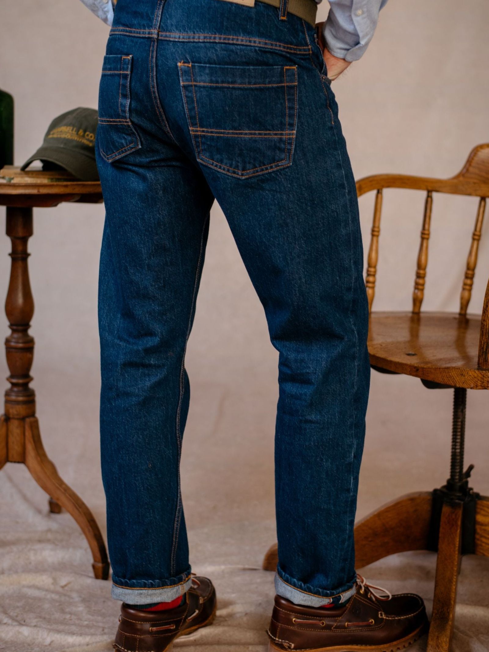 A person stands near a wooden chair and small table with a green cap, wearing Campbell's of Beauly Denim Jeans in medium-dark indigo selvedge and brown shoes; the photo, shot from behind, highlights these British-made jeans.