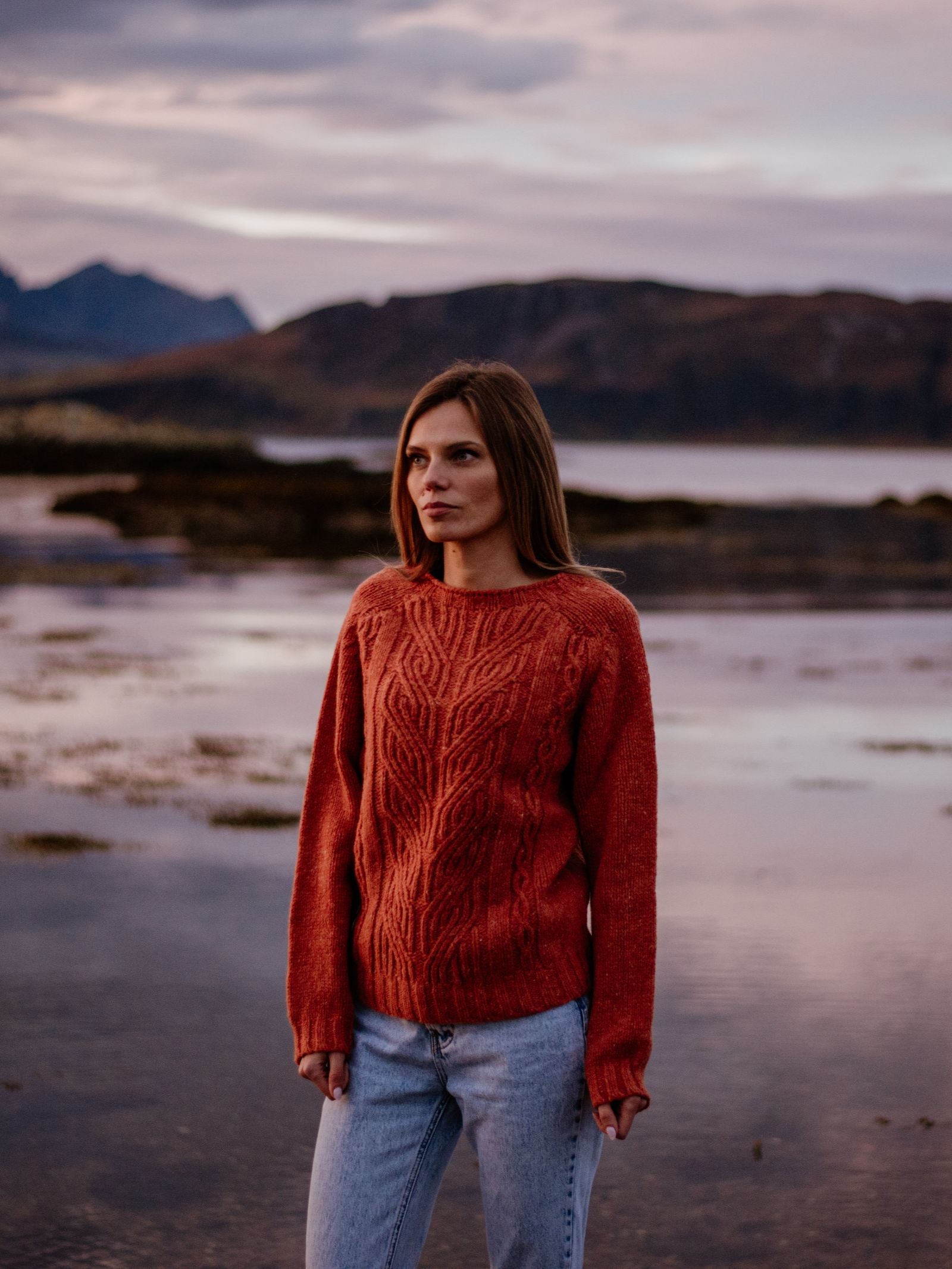 A woman in a rust-colored Auld Stock Donegal Lambswool Cable Jumper by Campbell's of Beauly and jeans stands by a calm lake with mountains behind her at dusk, beneath a cloudy sky.
