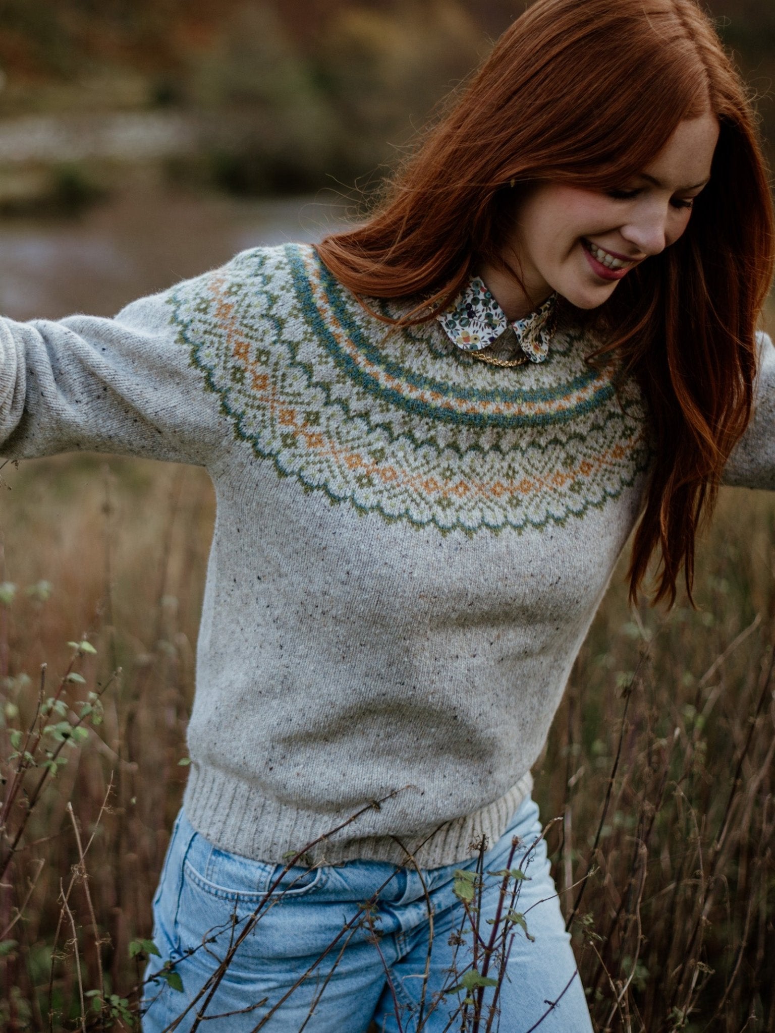 A young woman with long red hair smiles in a field, wearing Campbell's of Beauly's Auld Stock Donegal Lambswool Fairisle Crew Neck Jumper over a floral-collared shirt and light blue jeans.
