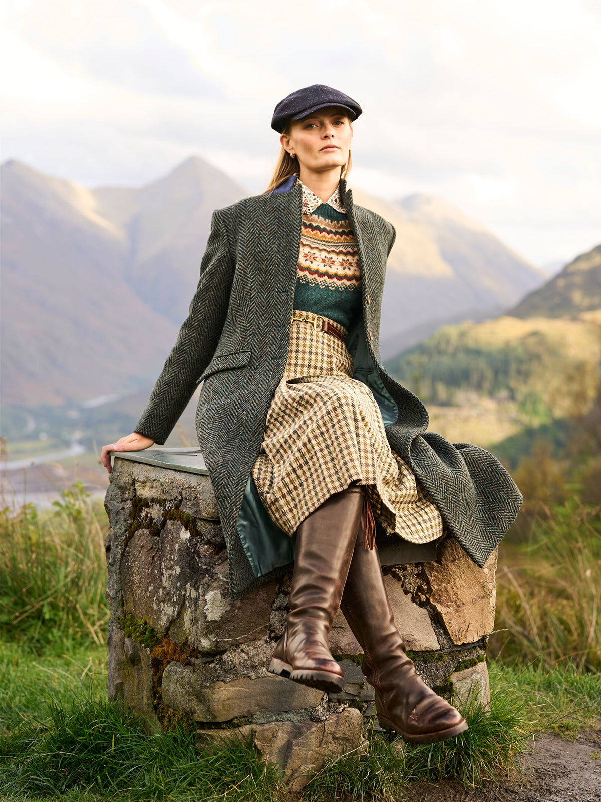 A woman in a flat cap, herringbone coat, patterned sweater, and a Campbell’s of Beauly Dogtooth skirt sits on a stone wall in a mountainous landscape.