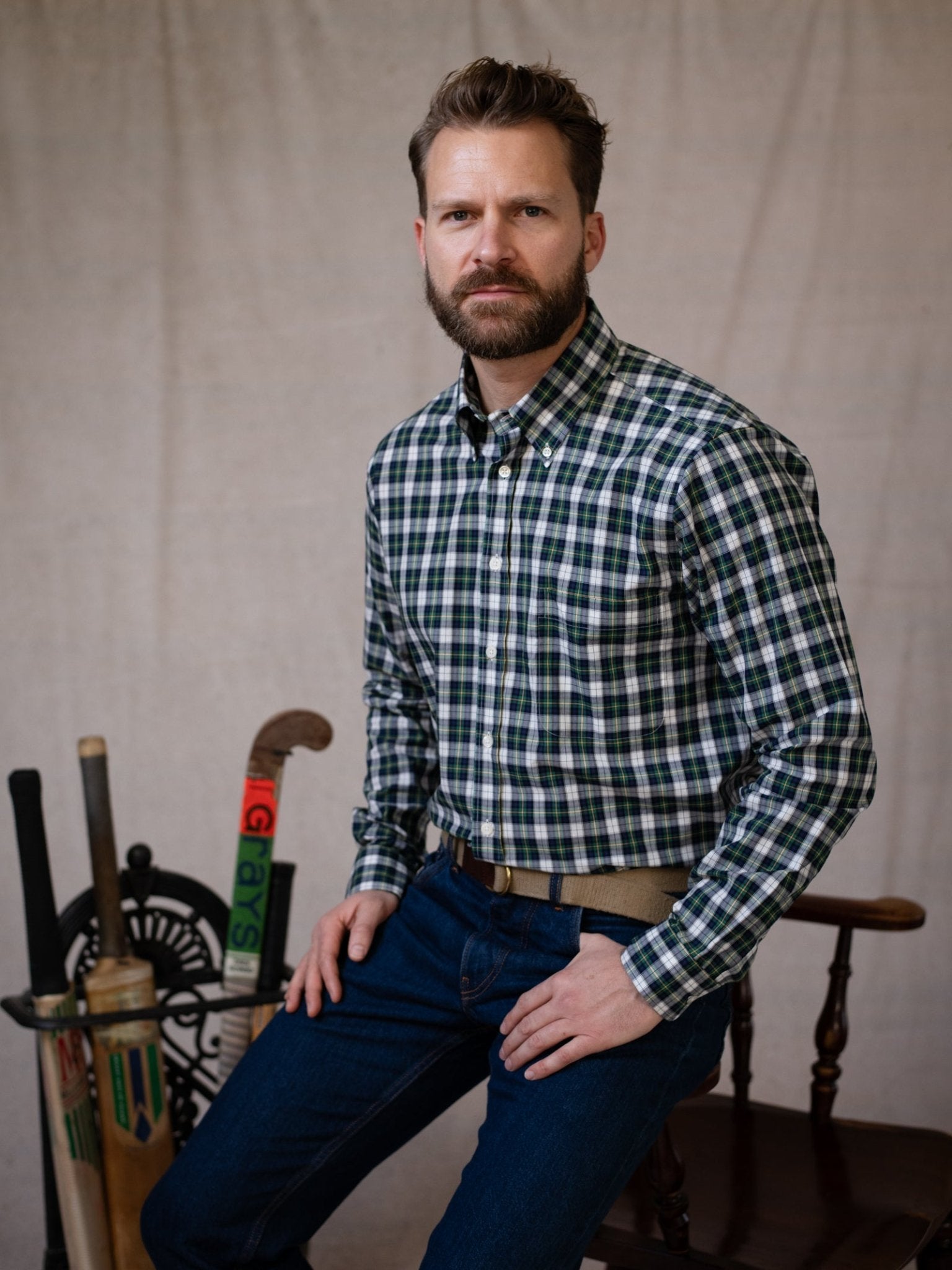 A bearded man wearing a Campbell's of Beauly Cotton Tartan Button Down Shirt in green and white sits on a chair, with vintage cricket equipment and a spinning wheel in the background.
