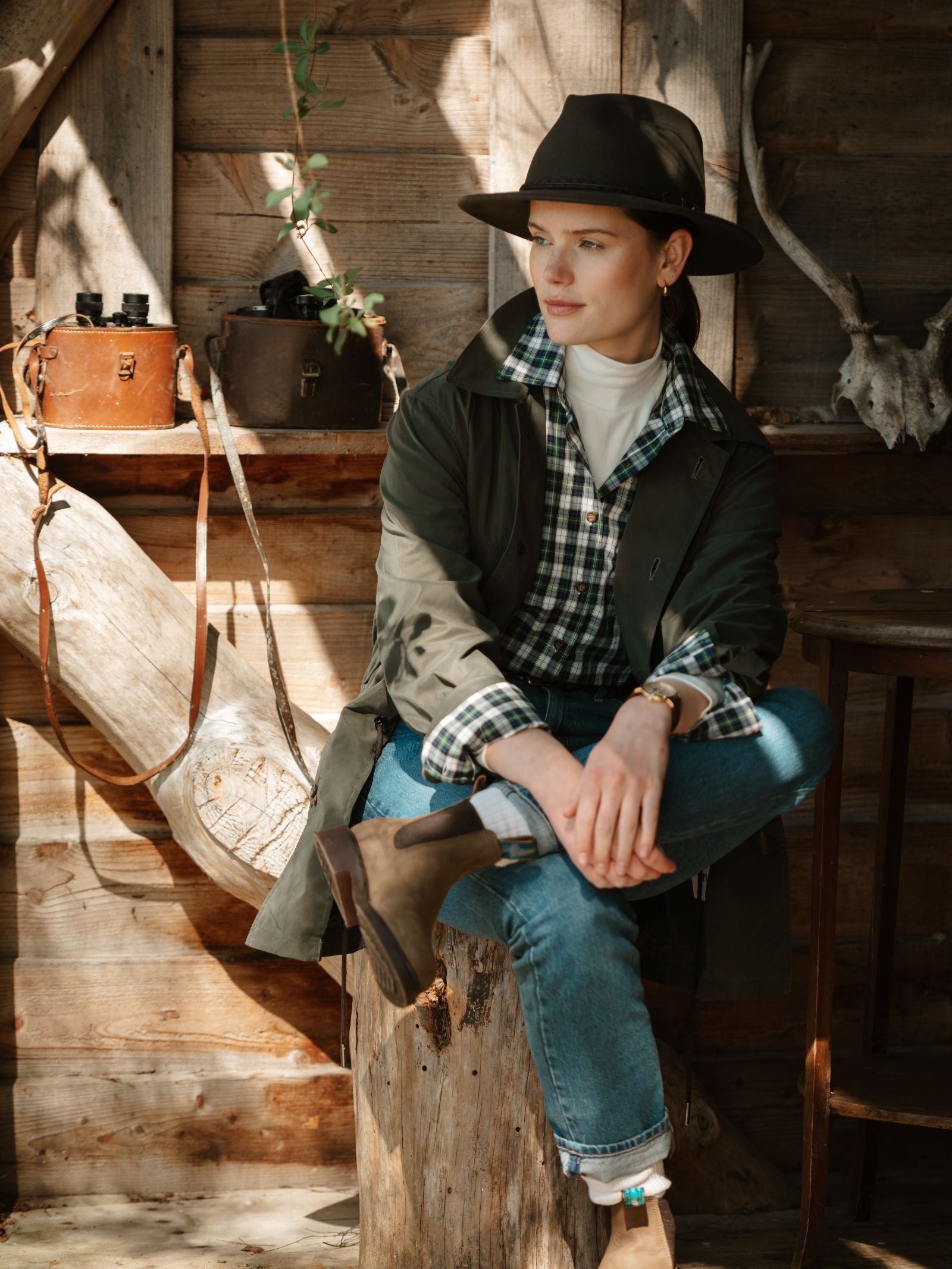 A woman wearing the Campbell's of Beauly Cotton Tartan Shirt in Dress Gordon sits on a tree stump in a rustic wooden cabin, sunlight streaming in over vintage leather bags and antlers for a warm, cozy atmosphere.