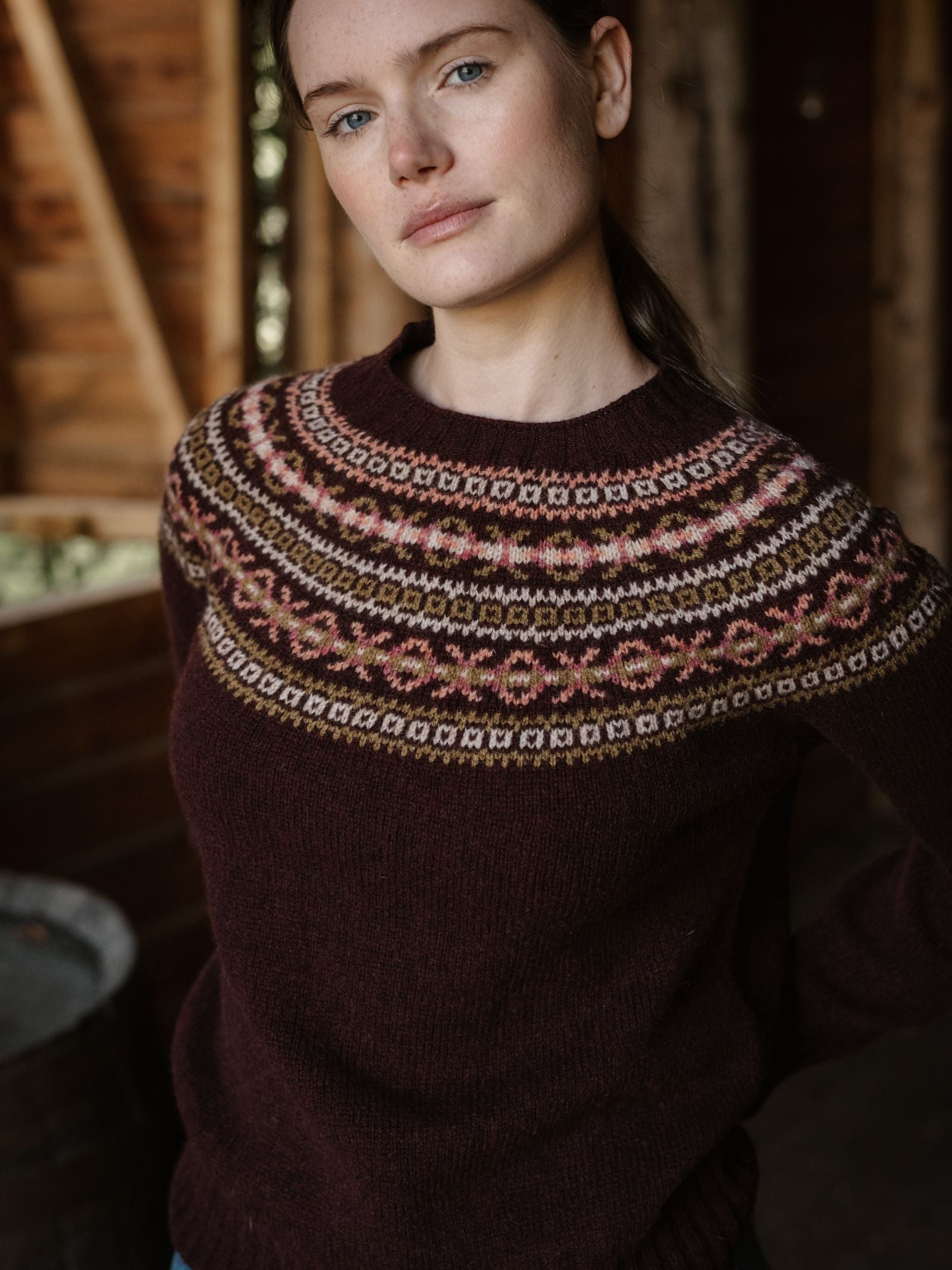 A woman with light skin and brown hair stands indoors, wearing a maroon Campbell's of Beauly Fairisle Crew Jumper. The cozy space features wooden walls and natural light. The jumper is knitted in Scotland from soft Geelong lambswool.