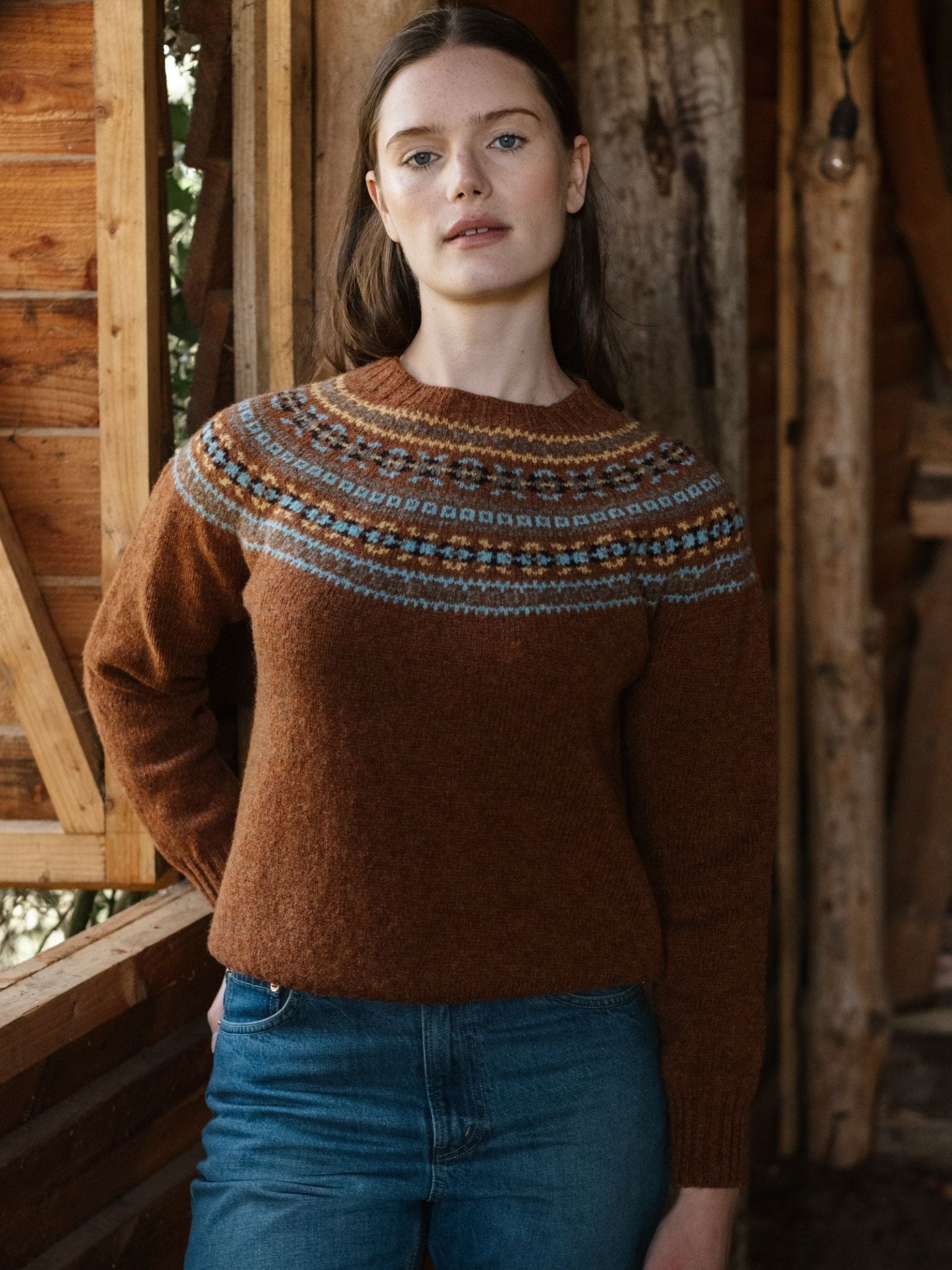 A woman with long brown hair stands indoors in a wooden cabin, wearing Campbell's of Beauly Fairisle Crew Jumper and blue jeans. She looks directly at the camera with a neutral expression.