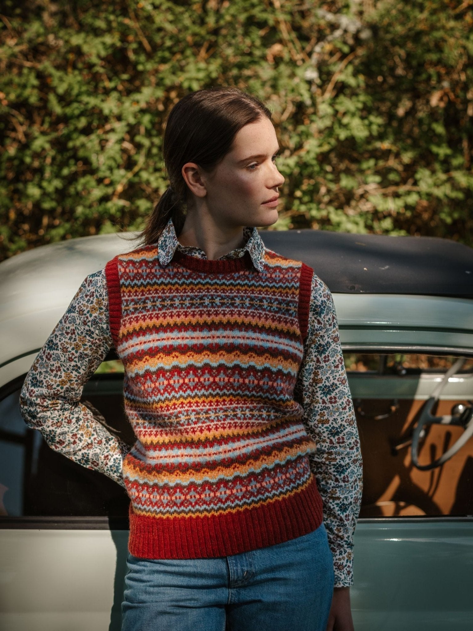 A woman wearing the Campbell’s of Beauly Fairisle Crew Tank, crafted in Scotland from fine Shetland wool, stands by a vintage car with trees behind her as sunlight casts gentle shadows and she looks to the side.