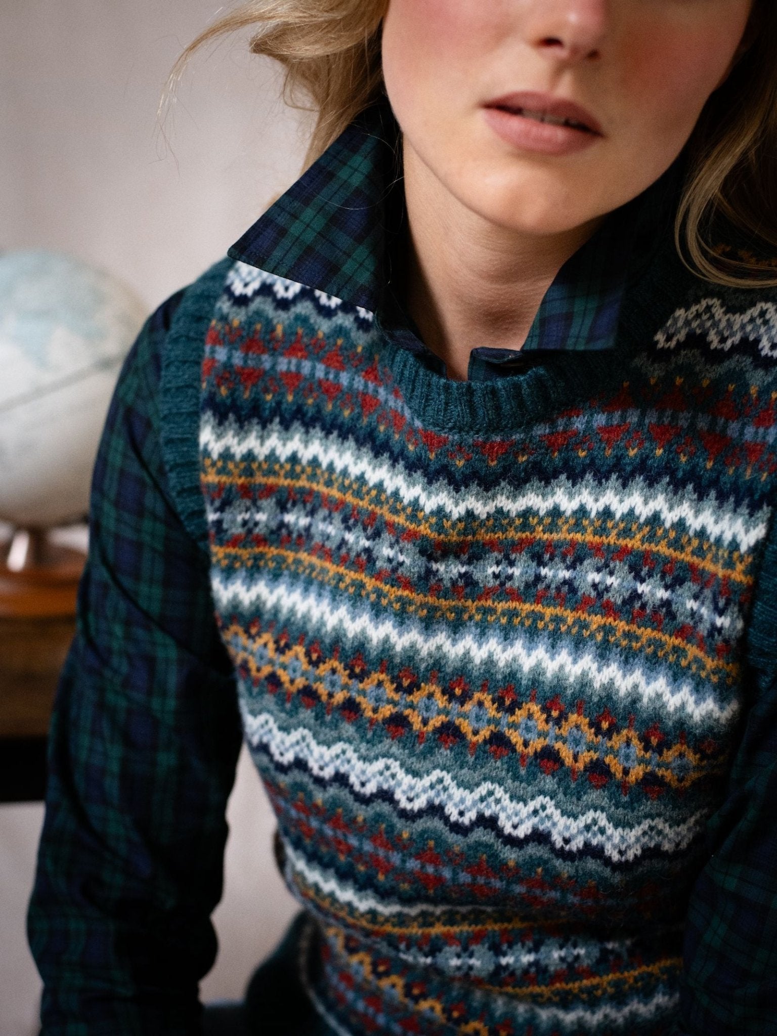 A person with loose hair and light pink lipstick sits beside a globe, wearing the Campbell's of Beauly Fairisle Crew Tank—featuring a blue, green, and orange Shetland wool knit—over a green plaid shirt. Only their upper body is visible.