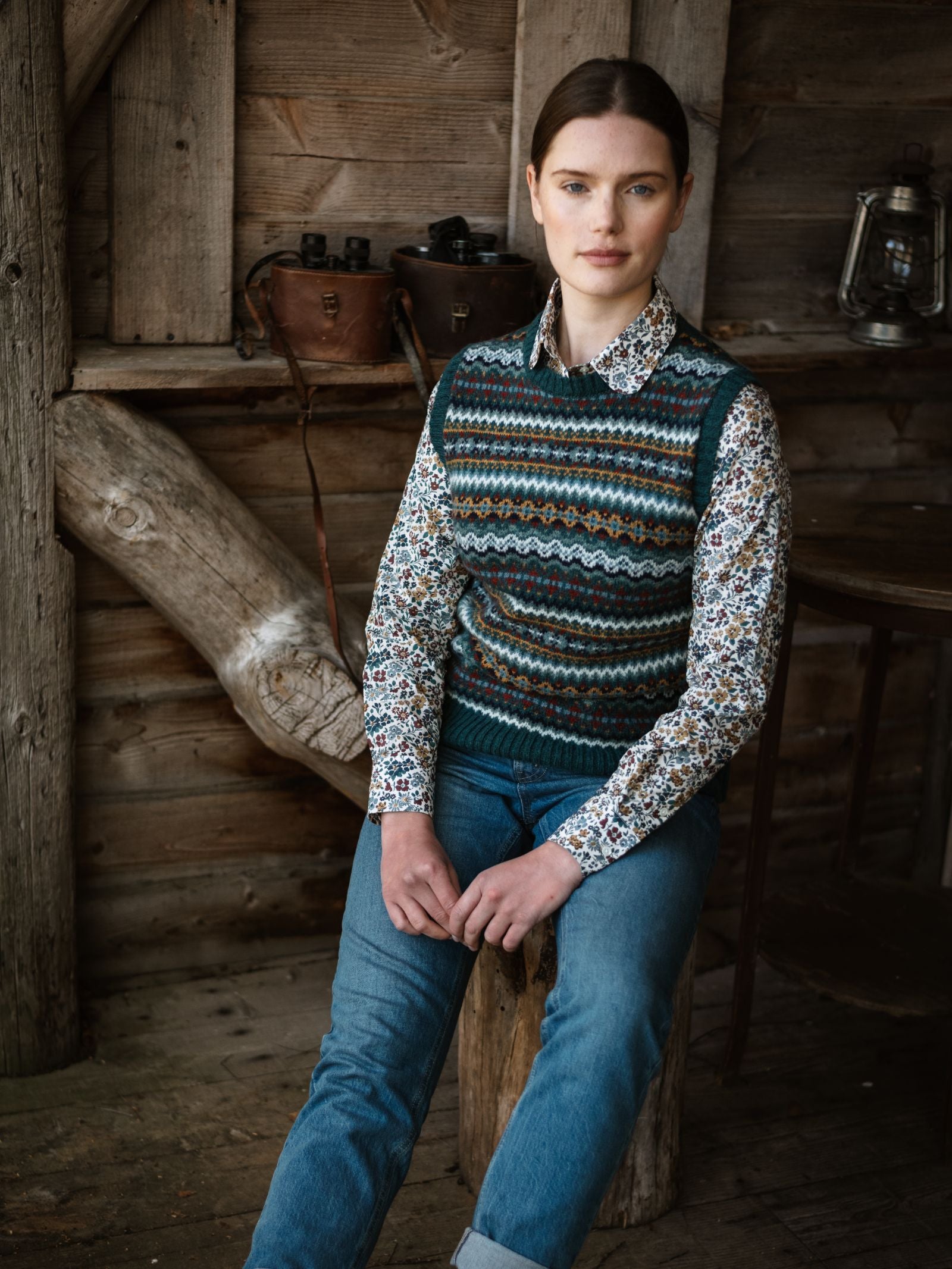 A person with brown hair sits calmly on a wooden stool in a rustic room, wearing blue jeans, a floral shirt, and the Campbell's of Beauly Fairisle Crew Tank, knitted in Scotland from soft Shetland wool.