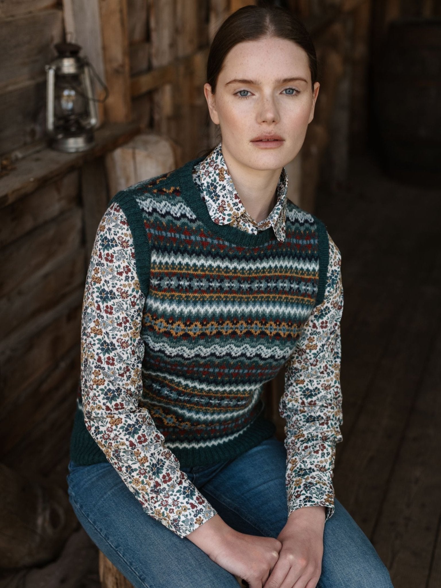 A woman with fair skin and brown hair sits indoors on a wooden bench, wearing the Campbell's of Beauly Fairisle Crew Tank over a floral shirt and blue jeans. A lantern hangs on the rustic wooden wall behind her.