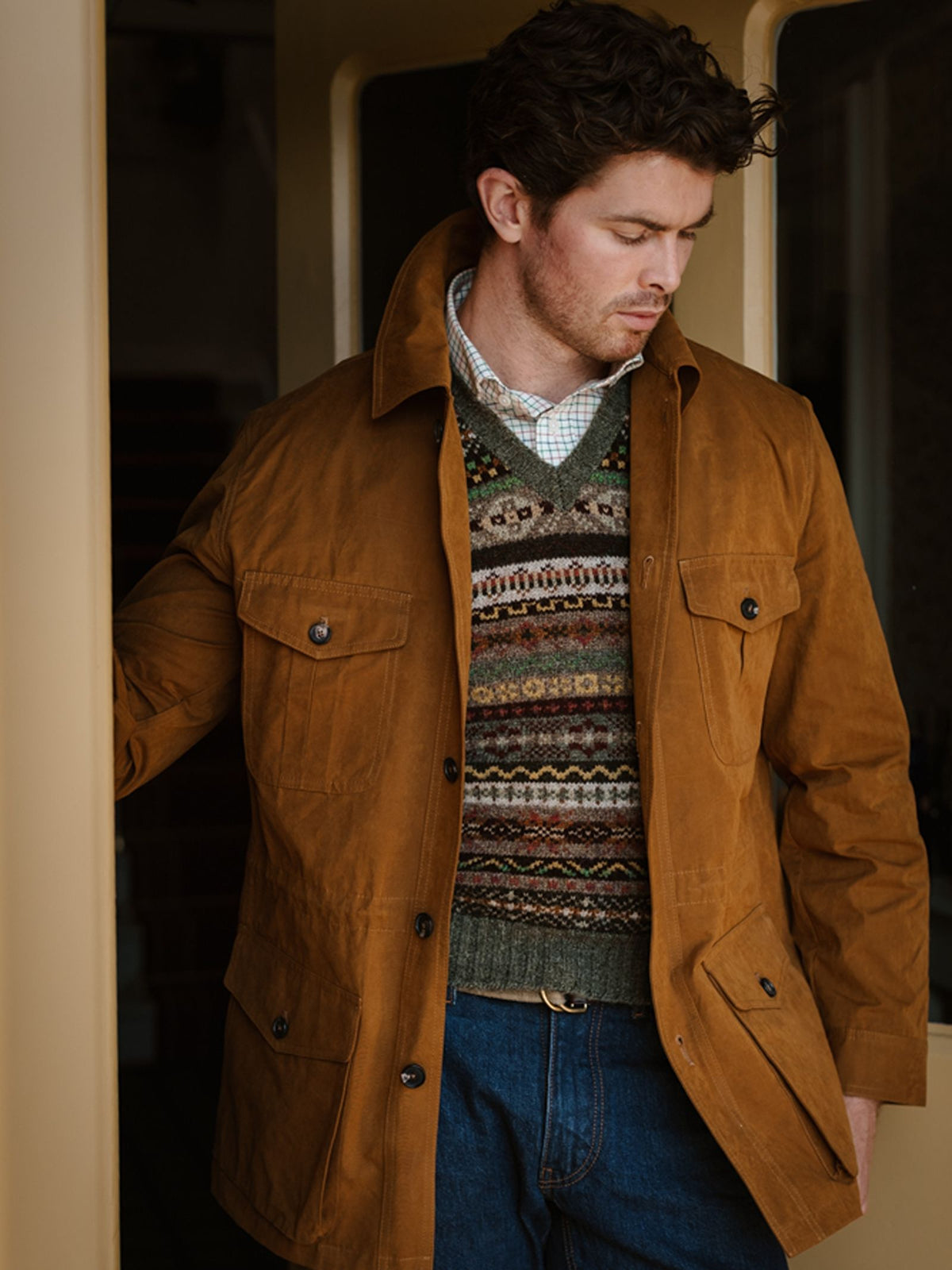 A man wearing a brown jacket, blue jeans, and the Campbell’s of Beauly Fairisle Slipover stands in a doorway, looking down with one hand resting on the doorframe.