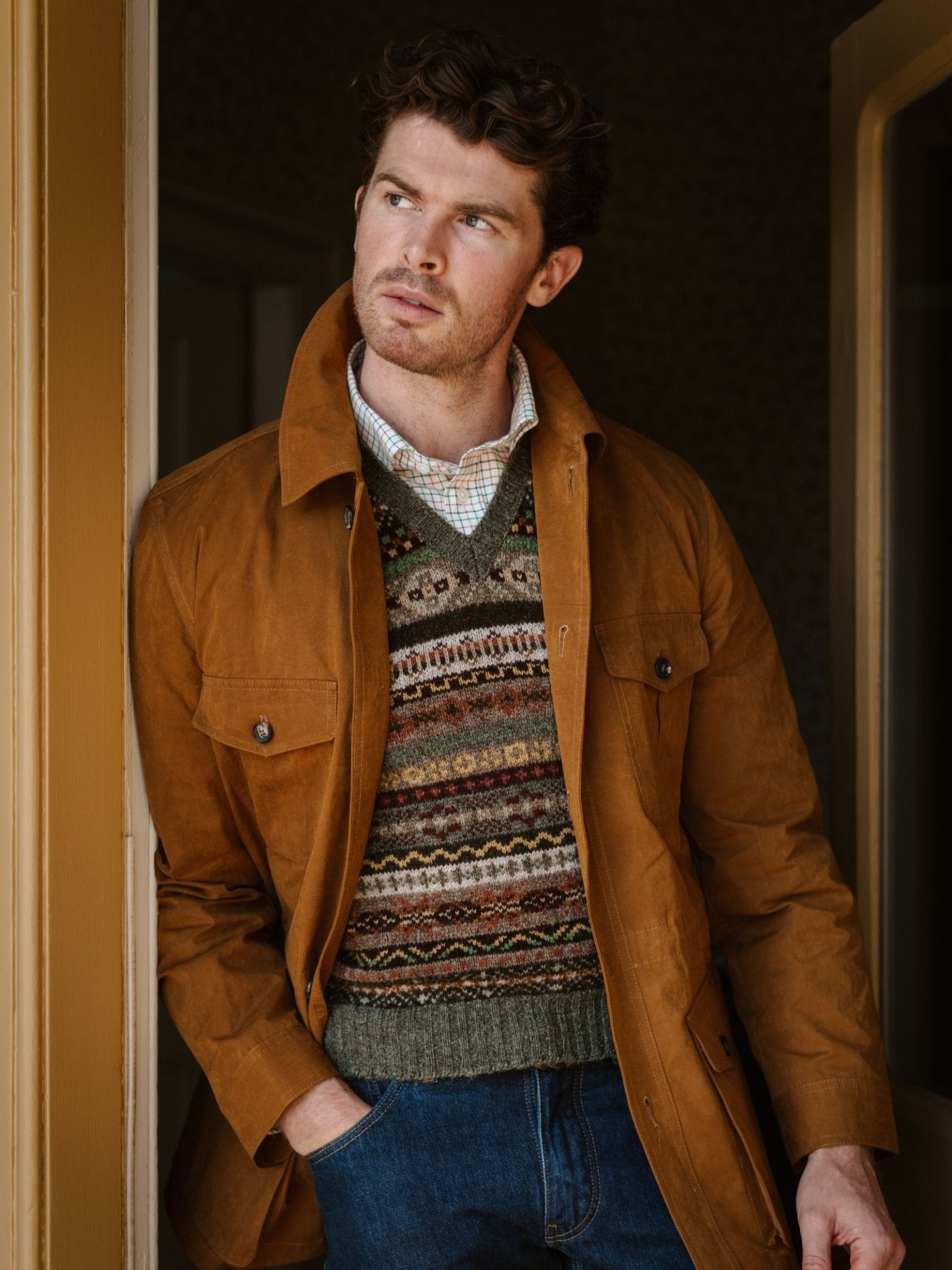 A man with curly brown hair wears a tan jacket over a Campbell's of Beauly Fairisle Slipover and checkered shirt. Indoors, he stands thoughtfully to the side with one hand in his jeans pocket.