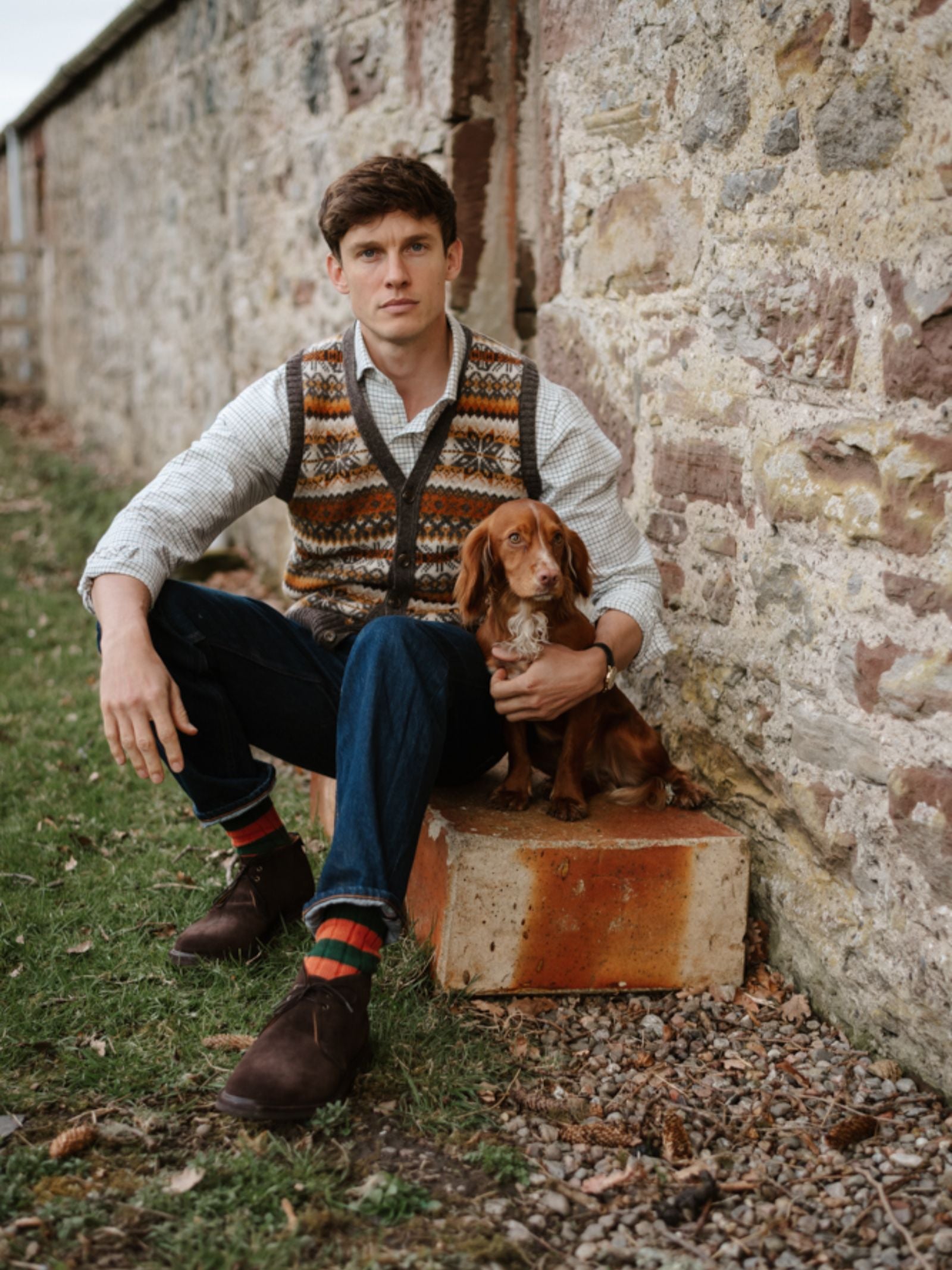 A man in a Campbell's of Beauly Fairisle V-Neck Waistcoat and jeans sits on a stone by a rustic wall, holding a brown dachshund on his lap outdoors on grass and gravel.