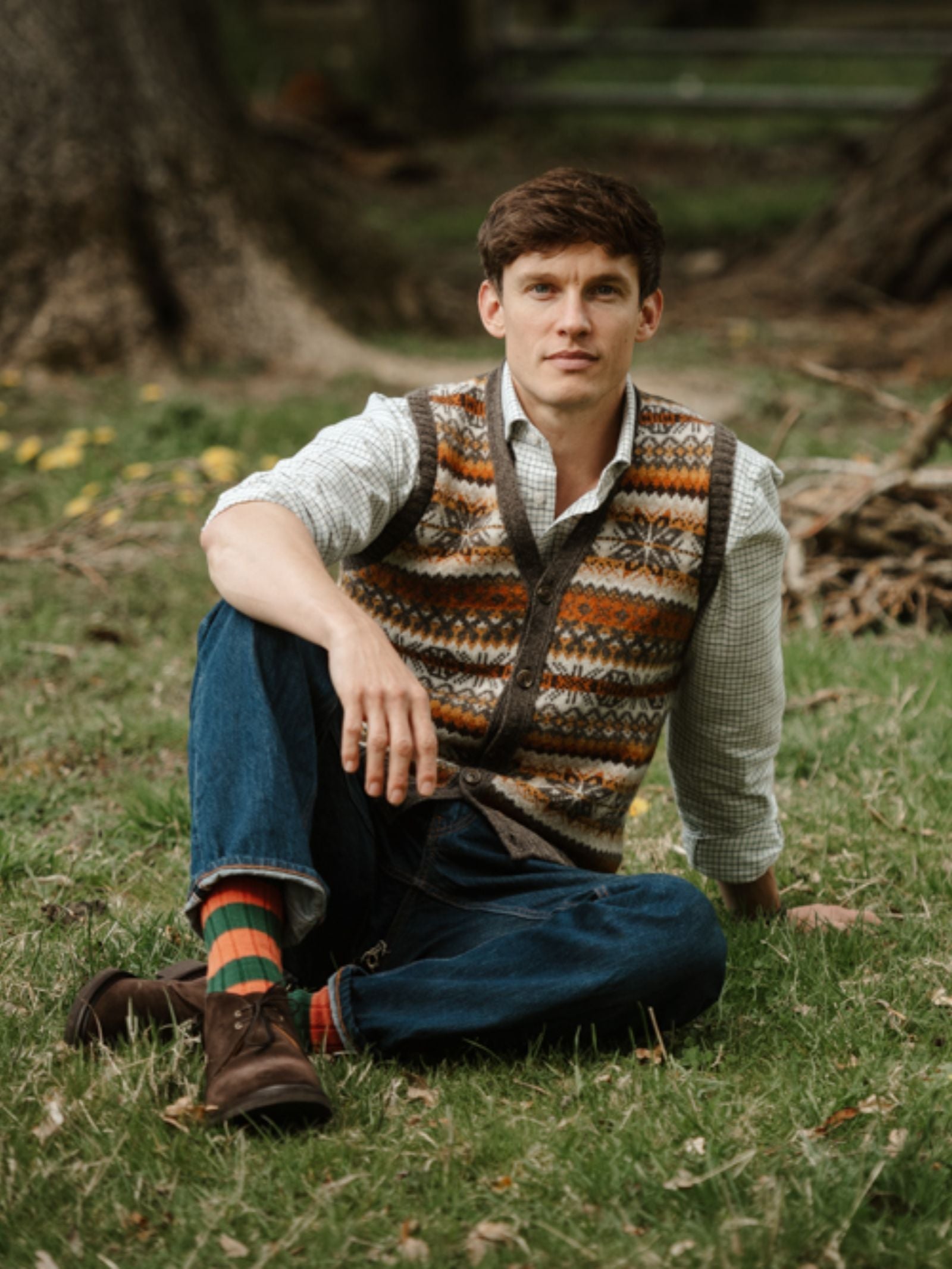 A young man relaxes outdoors on grass, wearing a Campbell's of Beauly Fairisle V-Neck Waistcoat in mocha over a plaid shirt, jeans, and brown boots. He looks at the camera, surrounded by trees and fallen branches.