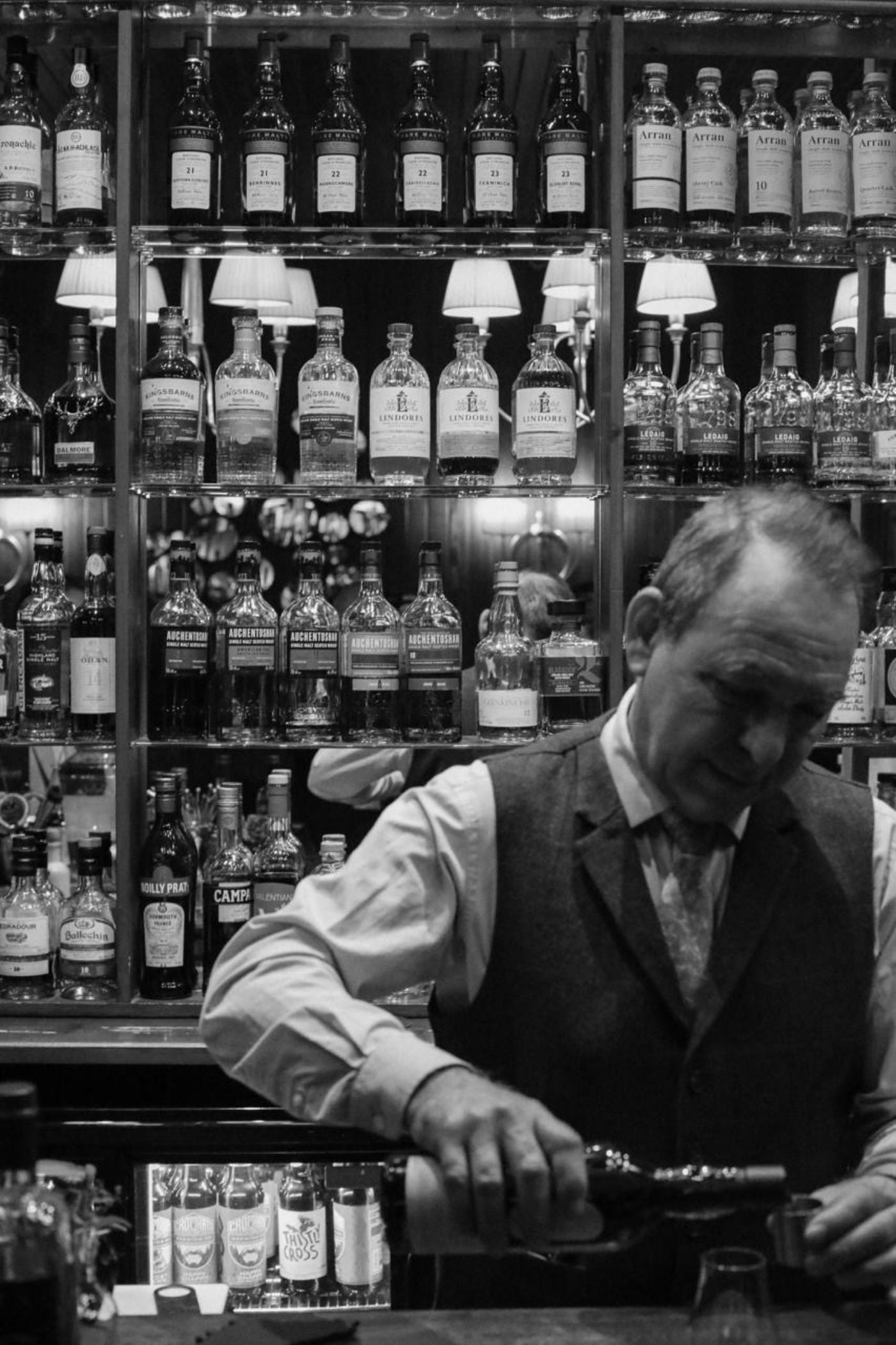 A bartender in a vest and tie pours a drink behind a bar, with shelves full of various liquor bottles in the background. The image is in black and white.