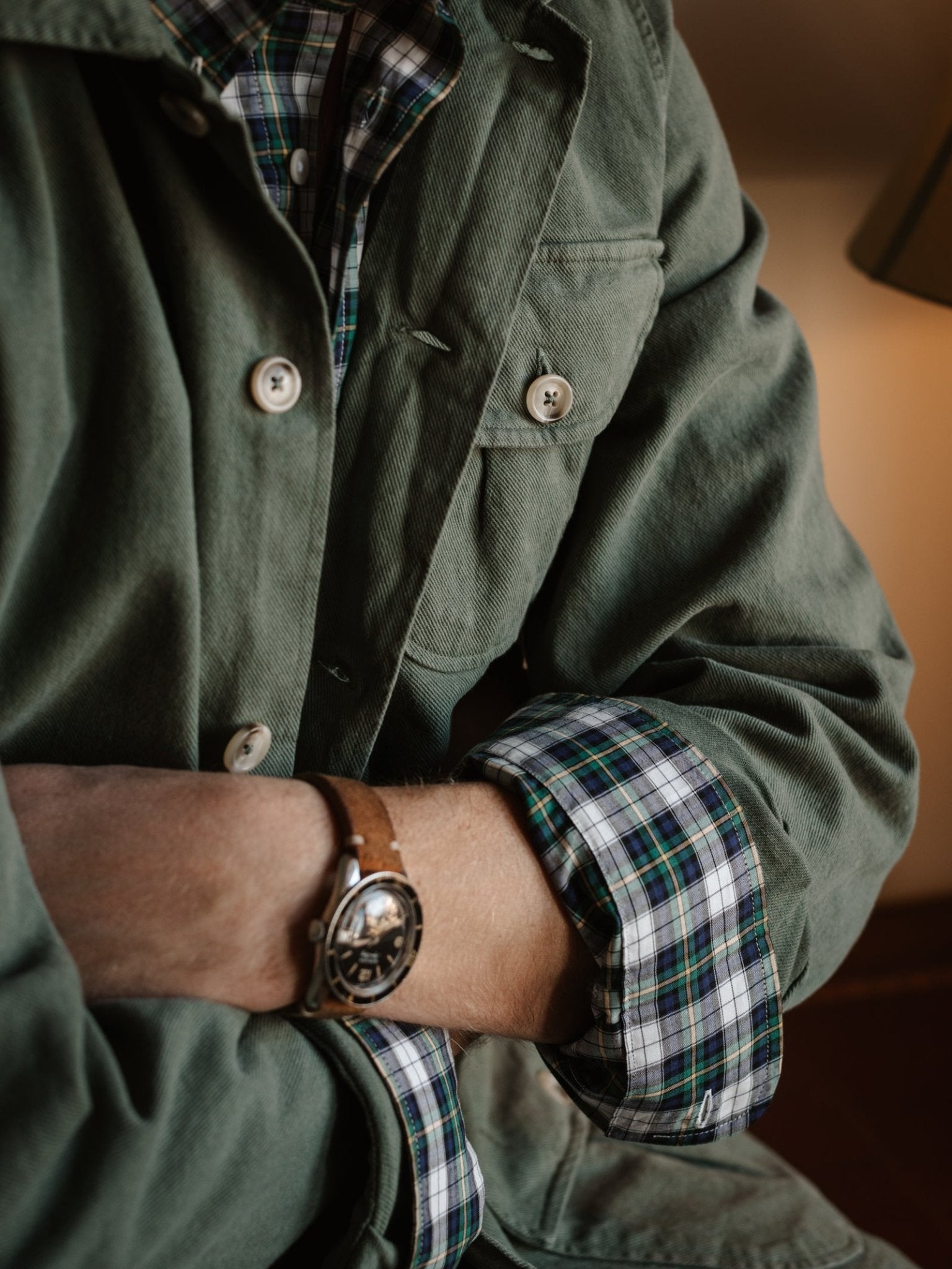 A person wears the Campbell's of Beauly Field Chore Jacket, crafted in Europe from 100% cotton, over a blue and green plaid shirt with rolled sleeves, revealing a brown leather strap wristwatch on their left wrist.