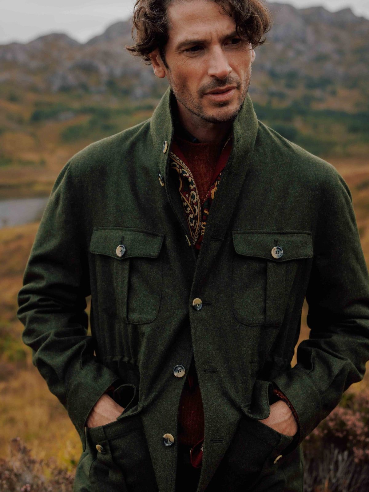 A man with wavy brown hair stands in a green field with hills behind him, wearing the Campbells of Beauly Field Teba Jacket over a red sweater and patterned scarf, his hands tucked into the jackets pockets.