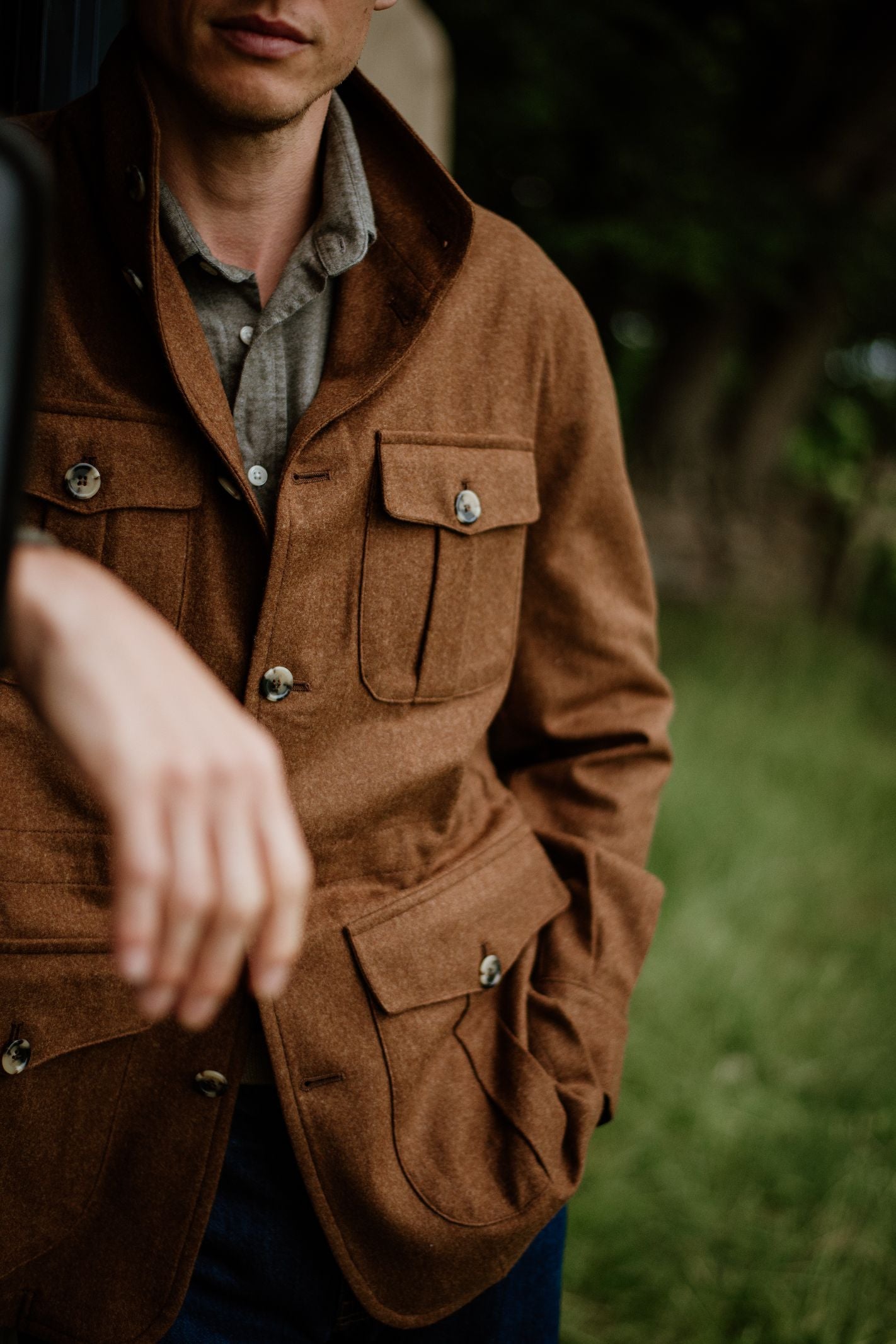A man wears the Campbell's of Beauly Field Teba Jacket with large pockets, standing outdoors in front of a green, blurred background. One hand is in his pocket while his other arm is extended forward.
