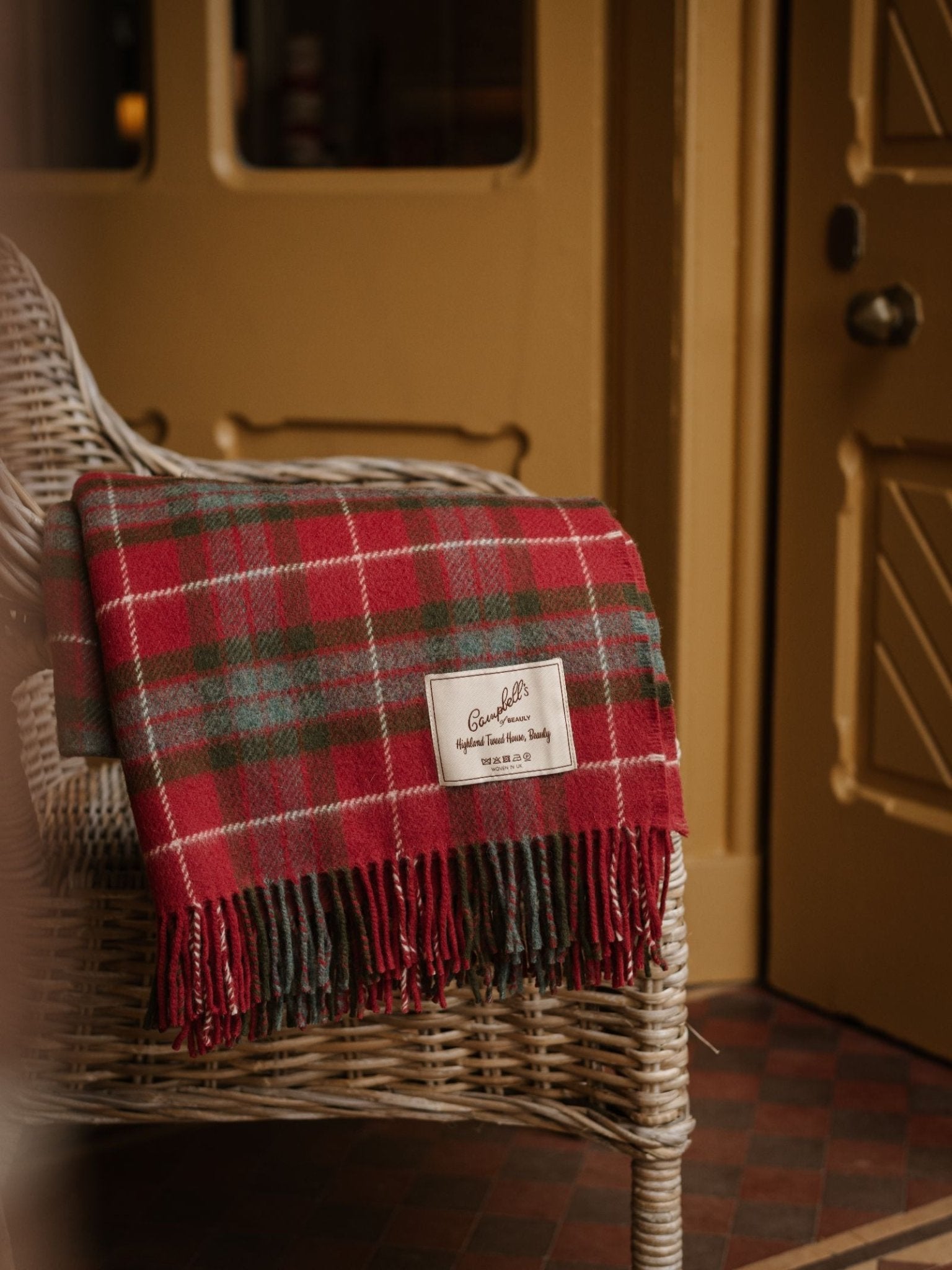 A Limited Edition Fraser Red Weathered Lambswool Throw by Campbell's of Beauly, in red and green plaid with fringe, is draped over a white wicker chair near a gold-colored door. The blanket features a "Genuine" label.