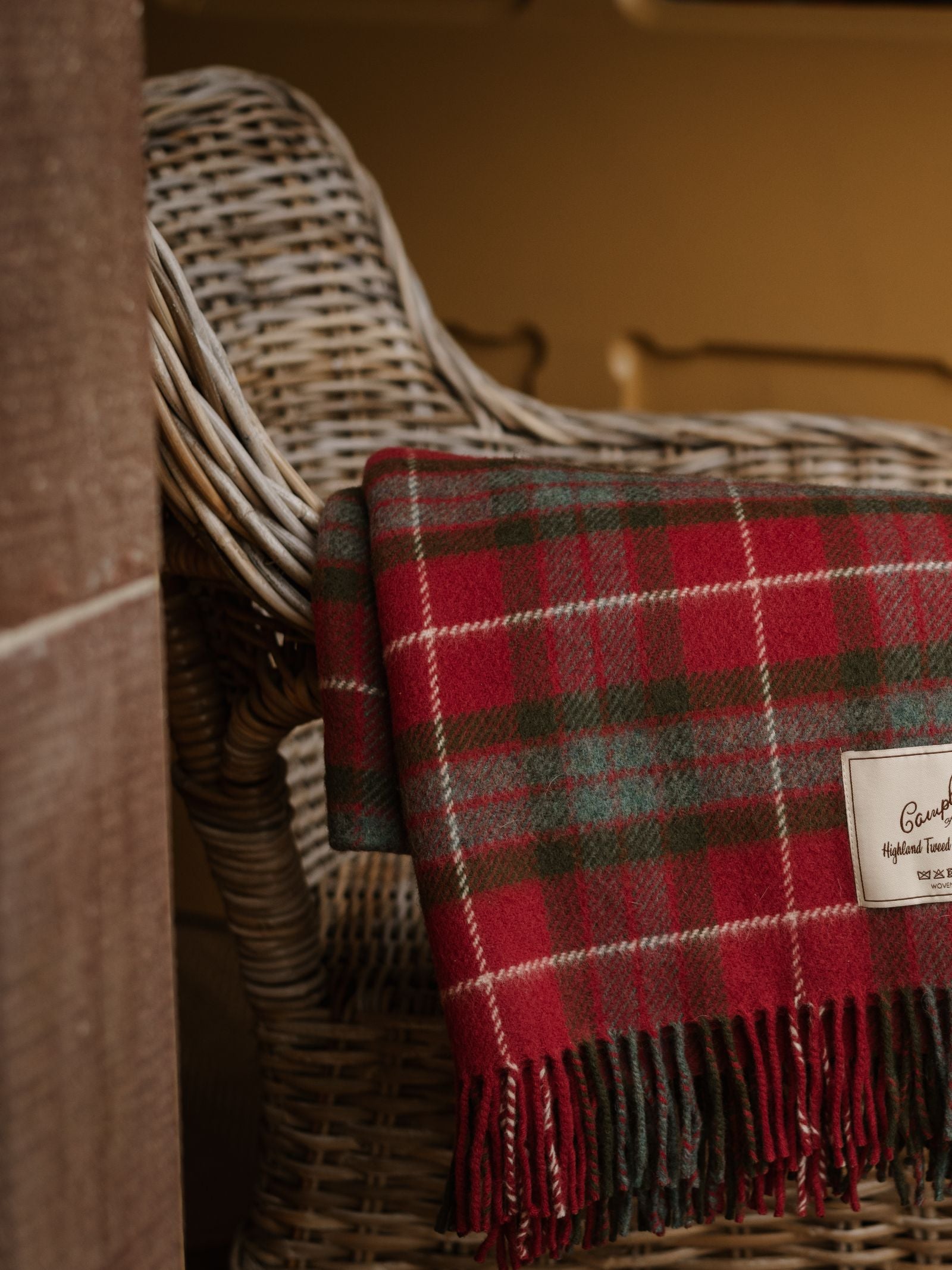 A Limited Edition Fraser Red Weathered Lambswool Throw from Campbell's of Beauly, featuring fringed edges, is draped over a wicker chair indoors, creating a warm and cozy atmosphere.