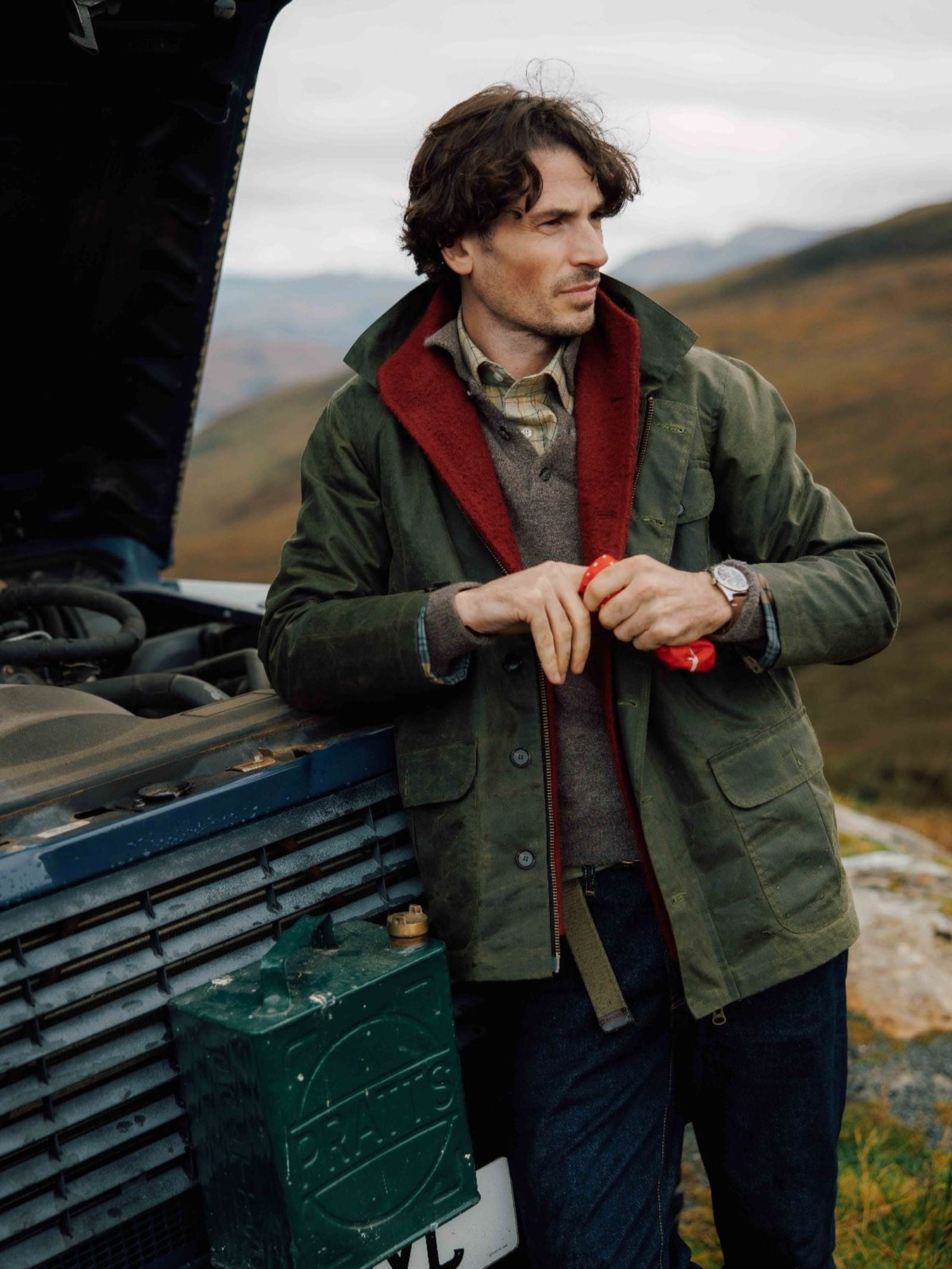 A man in the Campbell's of Beauly Garve Jacket leans against a vehicle with the hood open, holding a red cloth. He gazes thoughtfully over hill-covered terrain beneath cloudy skies—a perfect moment for this field-ready style.