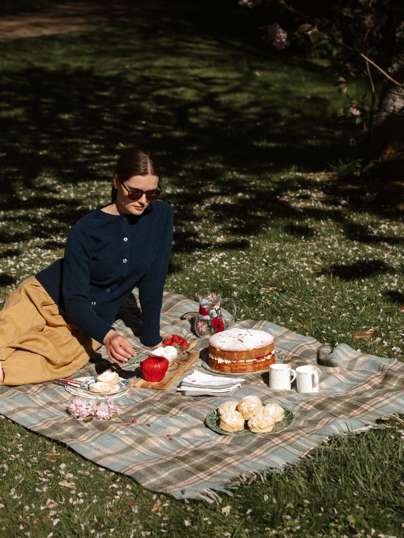 A woman in sunglasses and a navy Campbell's of Beauly Geelong Lambswool Crew Cardigan sits on a plaid blanket outdoors, enjoying a picnic with cake, pastries, mugs, and strawberries on a sunny lawn.