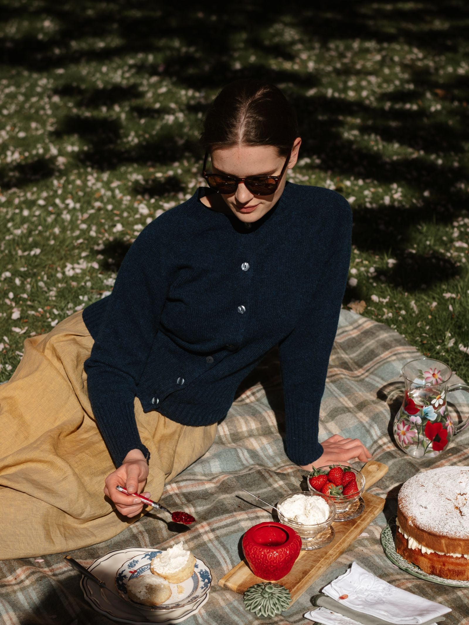Wearing sunglasses and a navy Geelong Lambswool Crew Cardigan by Campbell’s of Beauly, a woman sits outside on a plaid blanket, spreading jam on a scone amid sunlight, flowers, strawberries, cake, and a floral pitcher.