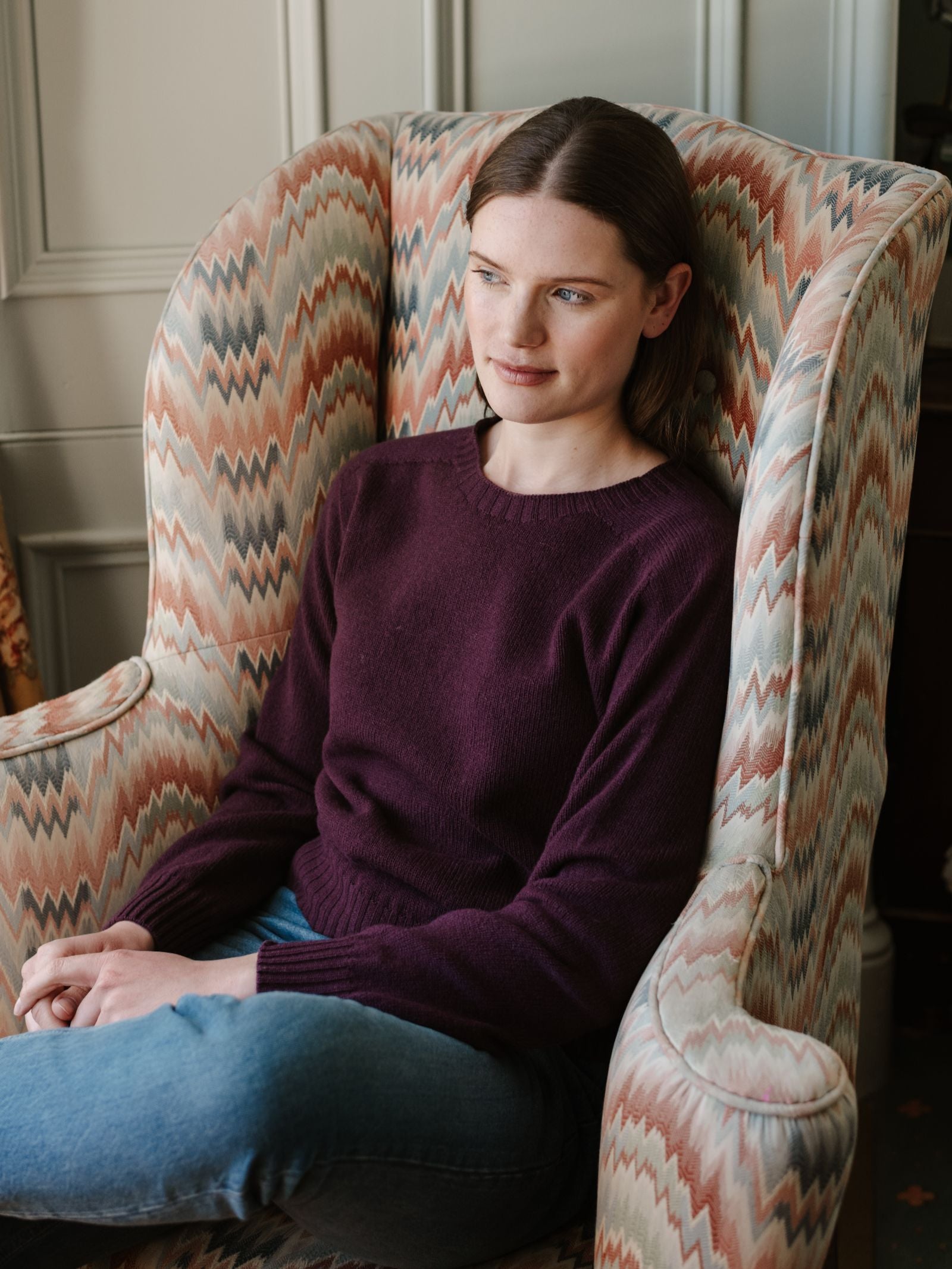 A woman with straight brown hair sits in a patterned armchair, wearing blue jeans and the Geelong Lambswool Crew Neck Jumper by Campbell's of Beauly. She looks relaxed, hands on her lap and legs crossed.