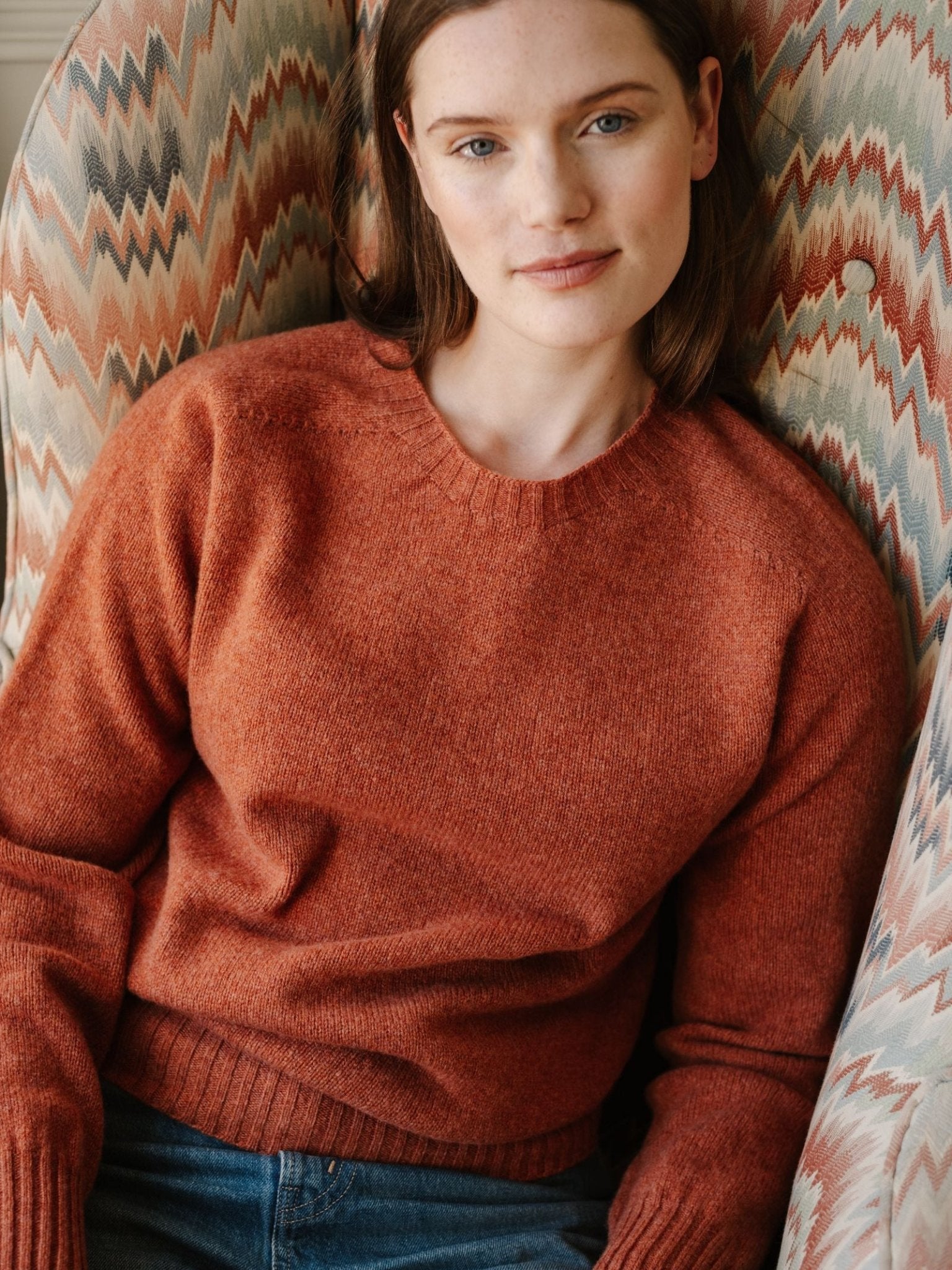 A woman with straight brown hair, wearing the Campbell's of Beauly Geelong Lambswool Crew Neck Jumper in rust and blue jeans, sits in a patterned armchair, softly posed against a wavy, multicolored fabric background.
