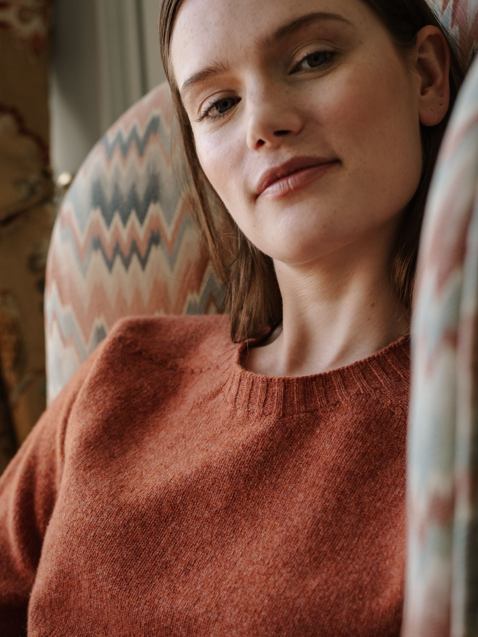 A woman with straight brown hair sits on a patterned chair, wearing the Campbell's of Beauly Geelong Lambswool Crew Neck Jumper in rust, and gazes softly at the camera with a relaxed expression.
