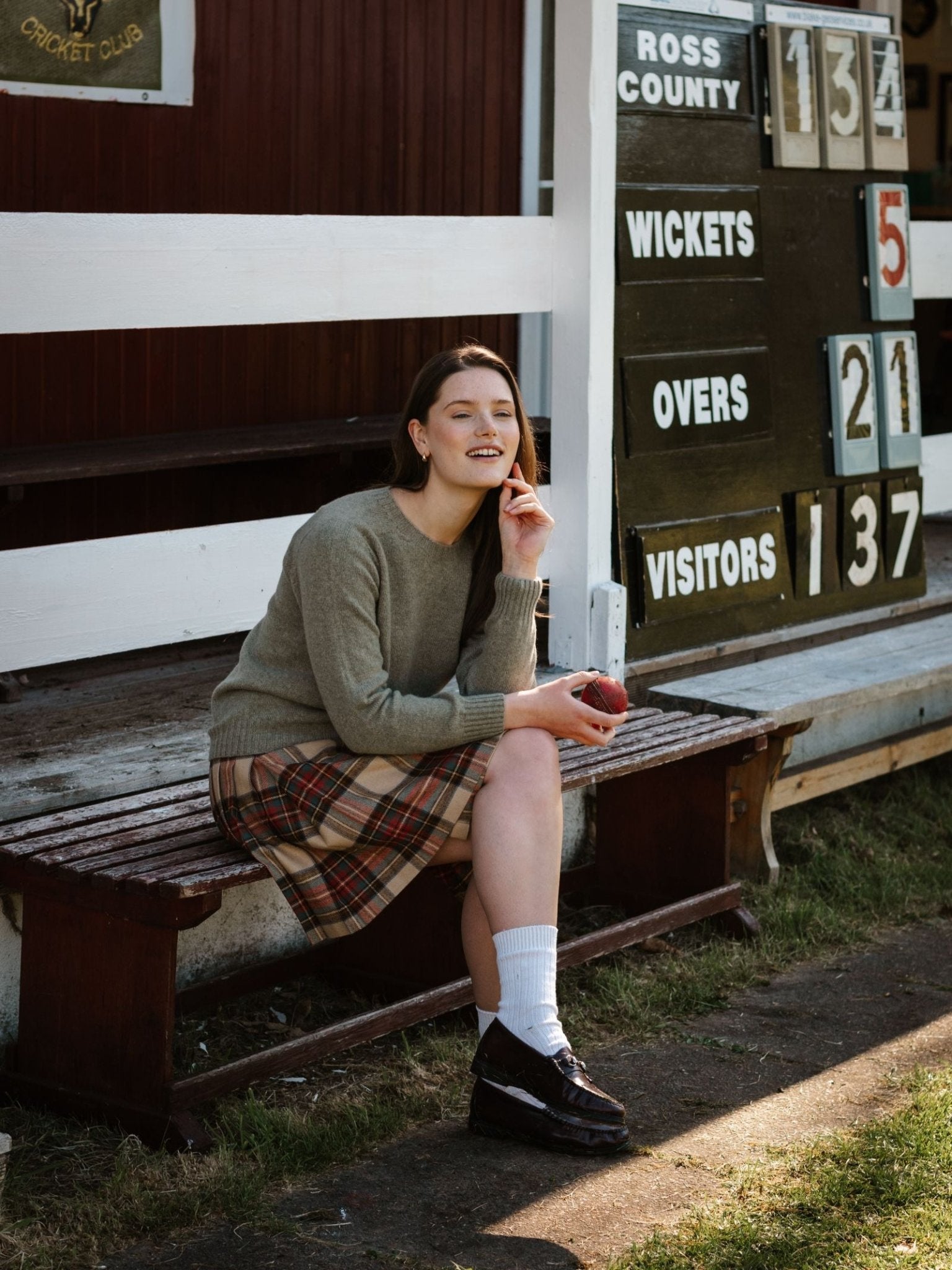 A young woman in a Campbell's of Beauly Geelong Lambswool Crew Neck Jumper and plaid skirt sits outdoors on a wooden bench with a cricket ball. Behind her, the scoreboard reads Ross County, 5 wickets down after 21 overs.