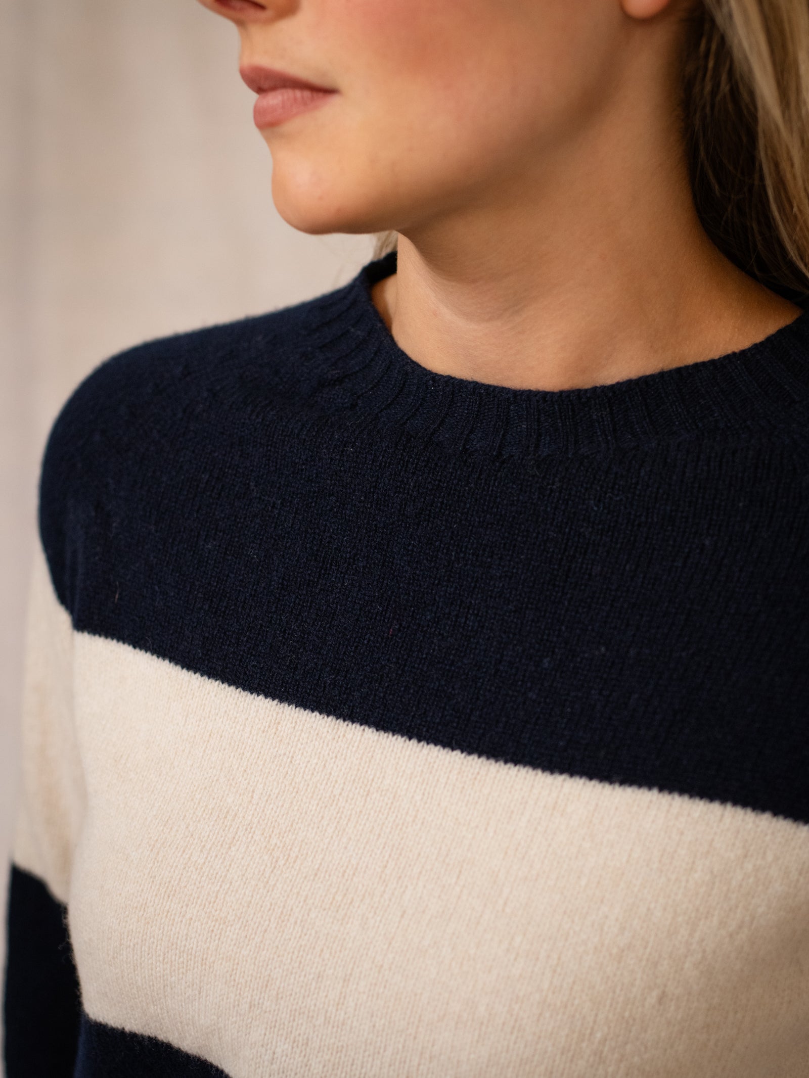 Close-up of a person wearing the Campbell's of Beauly Geelong Stripe Crew, a navy blue and white striped jumper knitted in Scotland from quality yarns, shown from chin down with part of the face and neck visible. Background is softly blurred.