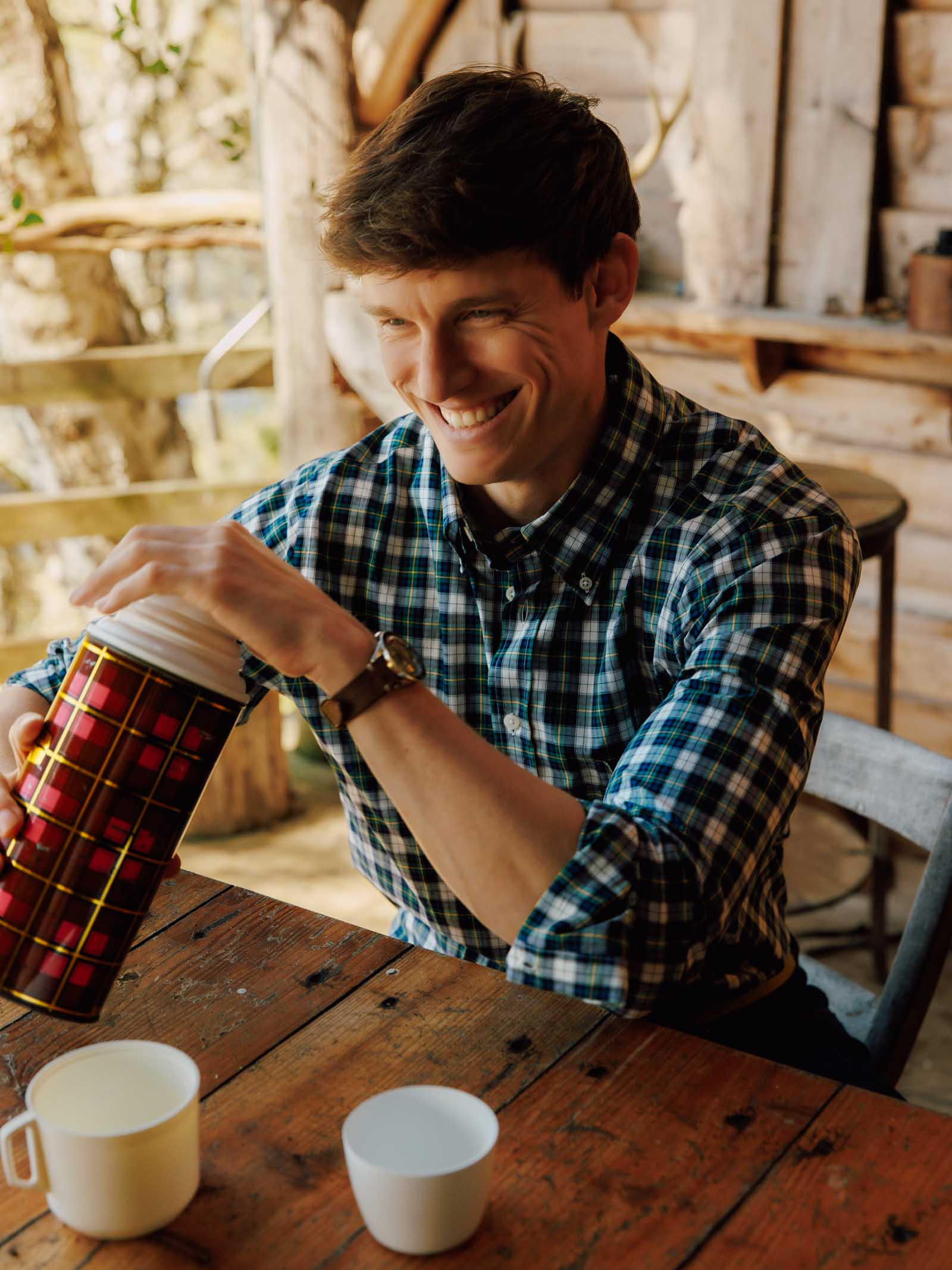 A smiling man wears a Campbell's of Beauly Cotton Tartan Button Down Shirt as he pours a drink from a red thermos into a white mug at a rustic wooden table, with a cozy cabin and trees in the background.