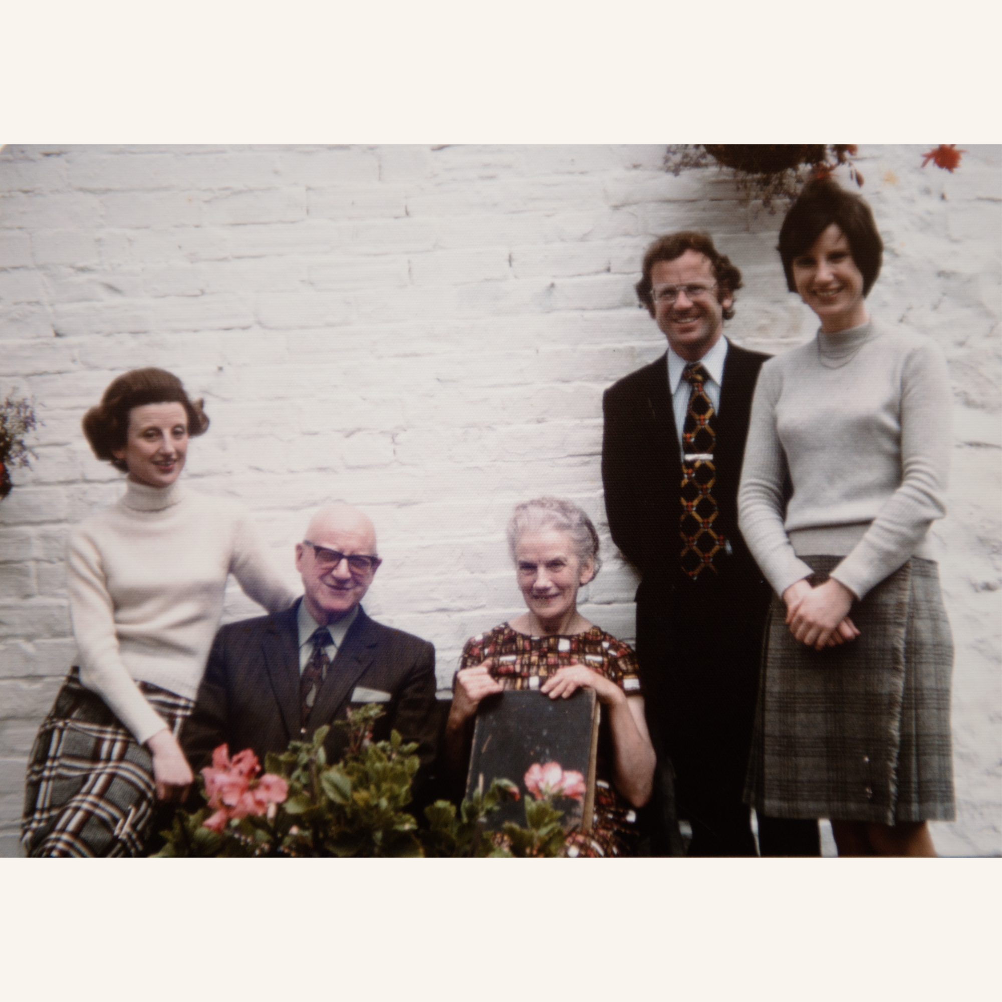 Five adults pose for a group photo in front of a white brick wall. Two men and three women, some seated and some standing, all dressed in vintage clothing, with flowers visible in the foreground.