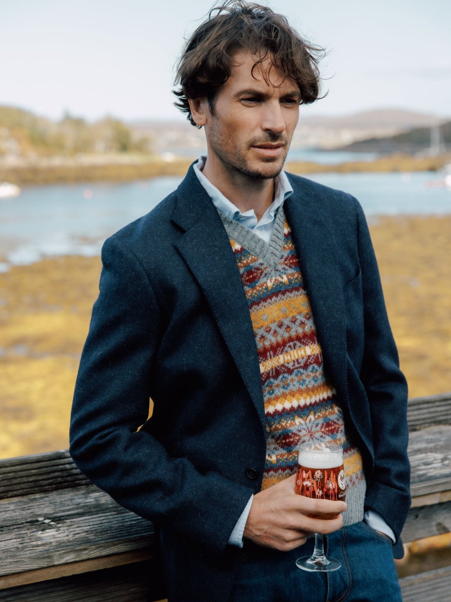 A man in a Campbell's of Beauly Campbell's House Jacket in navy twill stands by a wooden railing outdoors, holding a glass of beer with water and distant hills in the scenic background.