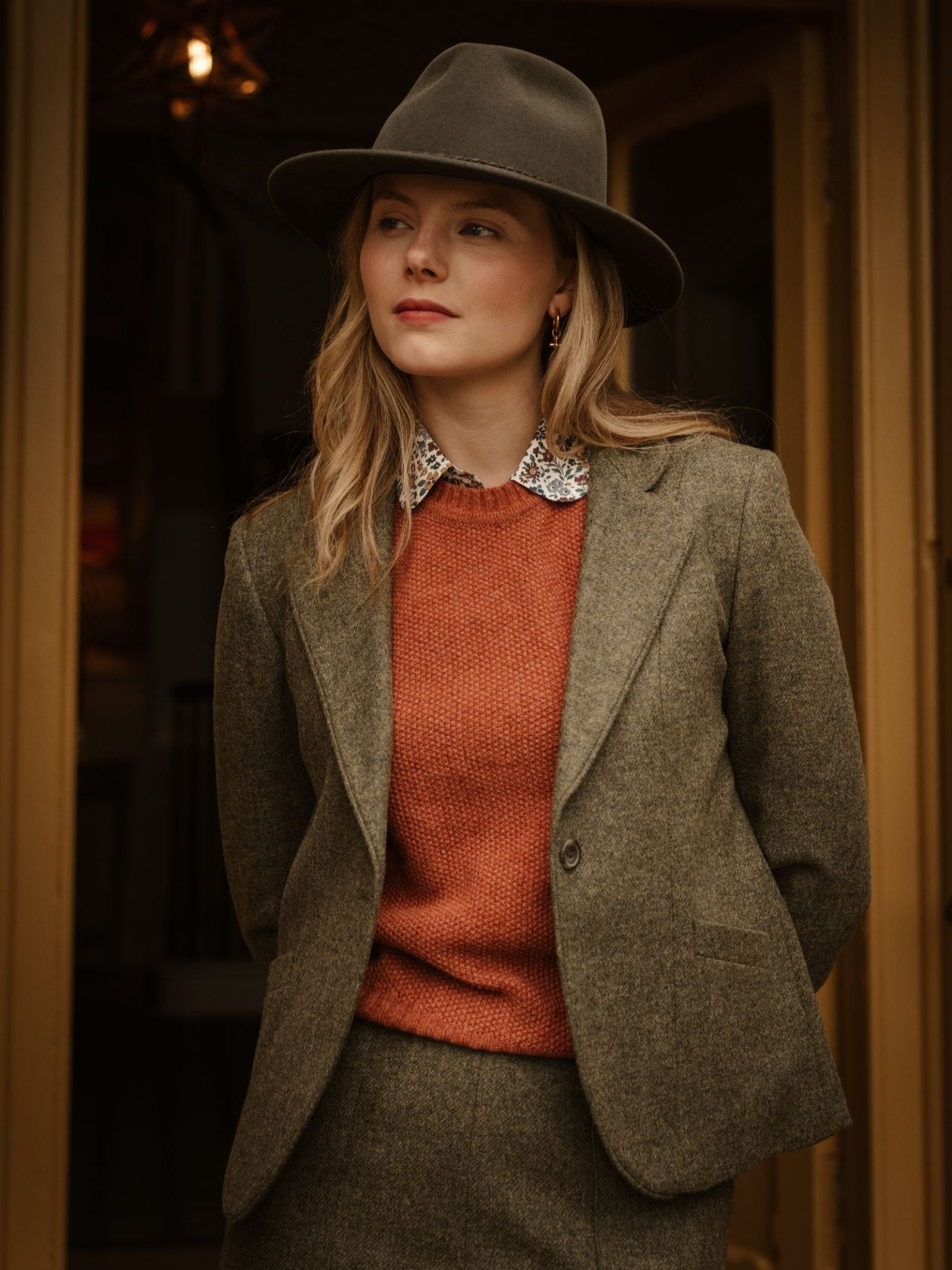A woman with blonde hair wears the Campbell's of Beauly House Jacket, an olive green skirt, orange sweater, patterned collared shirt, and a wide-brimmed hat as she stands outdoors looking to the side with a neutral expression.