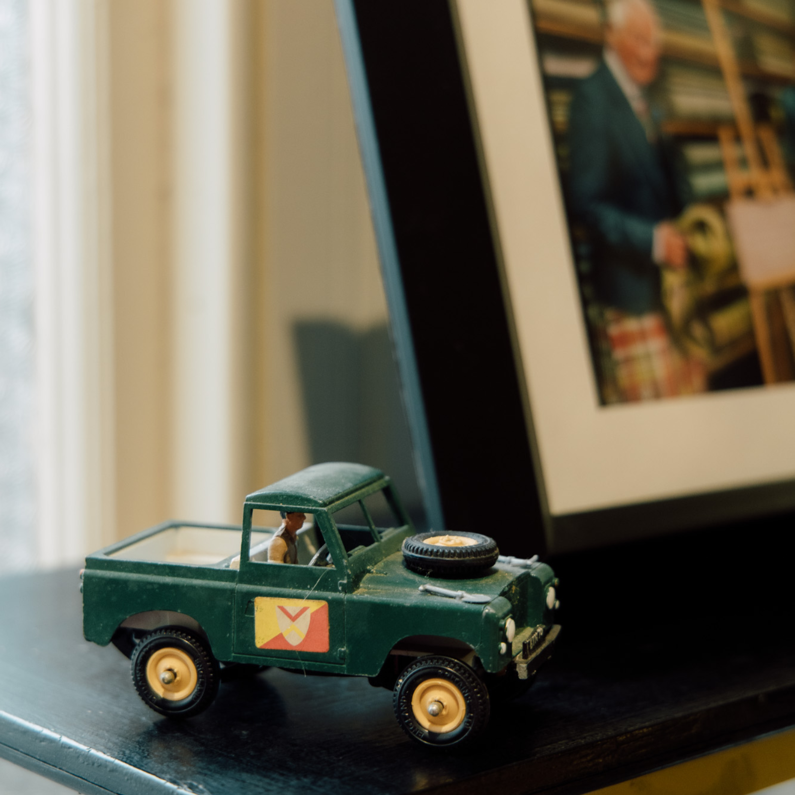 A small green toy truck with a yellow and red shield emblem is displayed on a dark surface next to a framed, blurred photo of a person indoors.