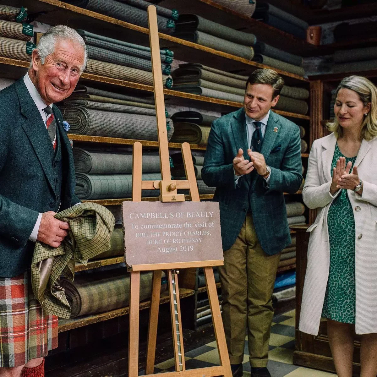 Prince Charles smiling and holding tartan fabric beside a commemorative plaque on an easel, with a man and a woman standing nearby clapping, surrounded by stacked rolls of fabric.