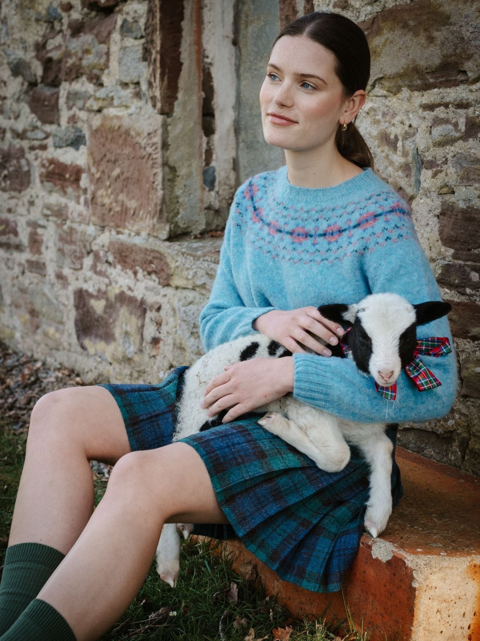A woman wearing a blue patterned sweater and Campbell's of Beauly Tweed Kilt sits on a stone step by a rustic wall, holding a black-and-white lamb adorned with a red tartan bow.