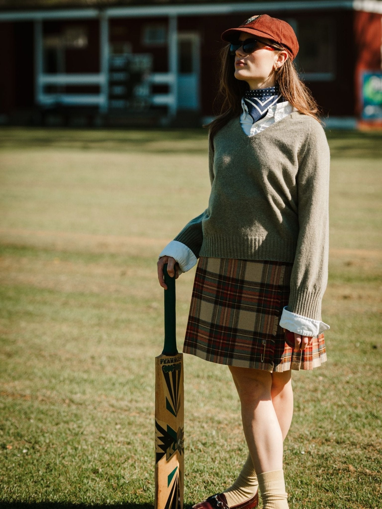 A woman wearing a Campbell's of Beauly Tweed Kilt, sweater, collared shirt, cap, sunglasses, and scarf stands on grass holding a cricket bat, with a red building in the background.