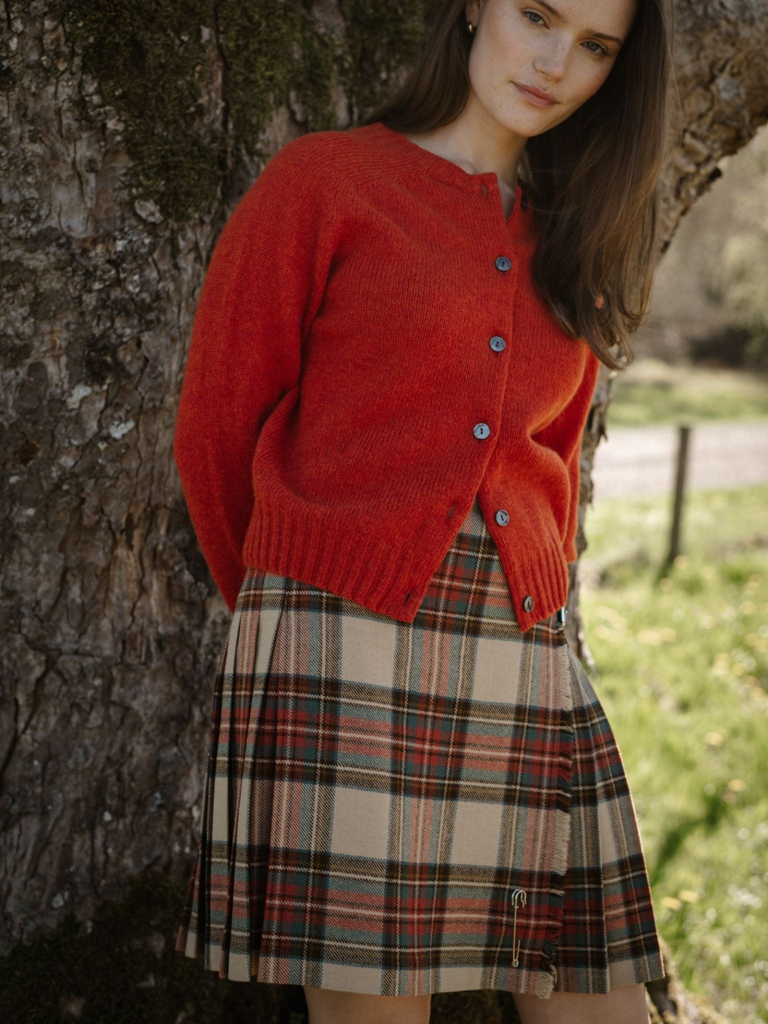 A woman stands outdoors by a tree, wearing a Campbell's of Beauly Tweed Kilt and a bright red buttoned cardigan. She has long brown hair and a calm expression as sunlight filters through the background.