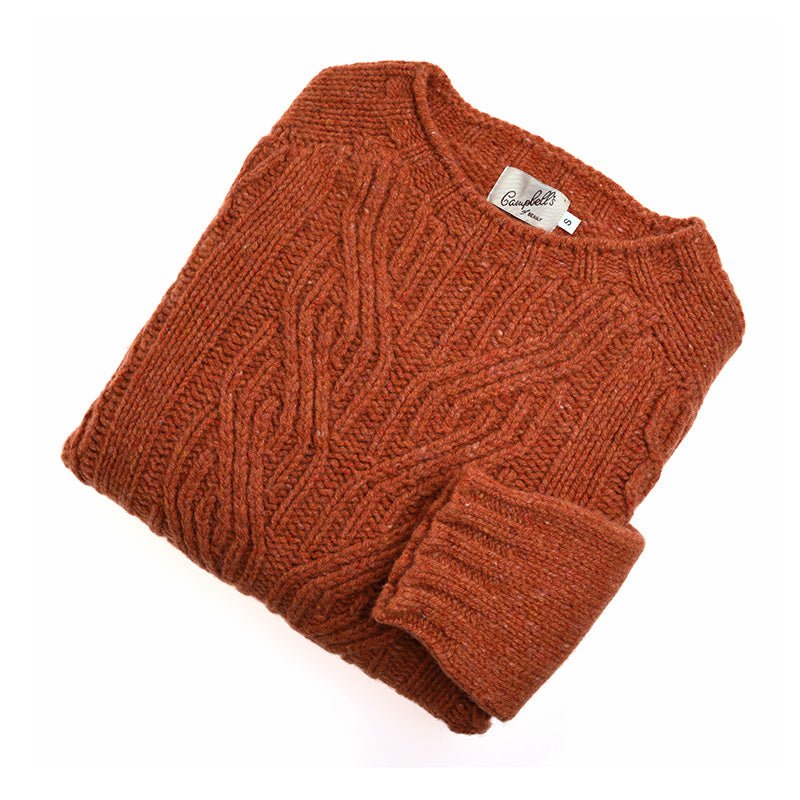 The Campbell's of Beauly Auld Stock Donegal Lambswool Cable Jumper is a chunky, rust-orange textured sweater with a cable knit pattern. It’s shown folded on a white background, featuring a visible tag inside the collar.