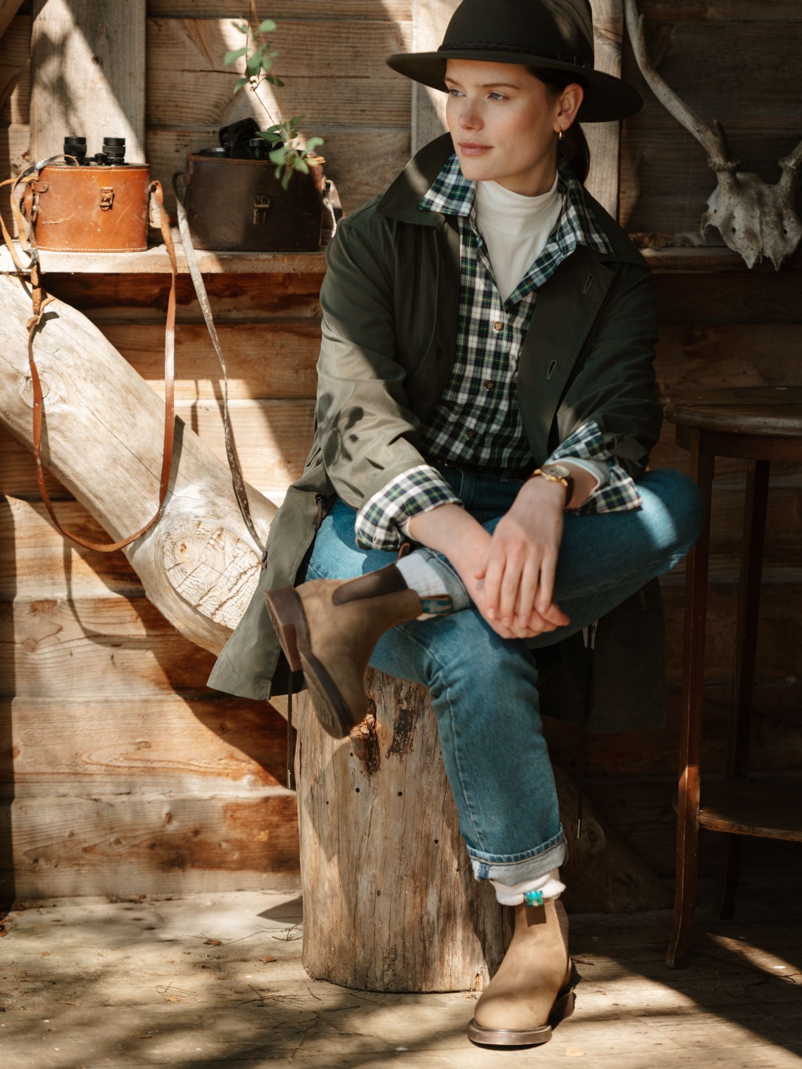 A person in a wide-brim hat, green coat, plaid shirt, jeans, and the Ladies' R.M.Williams Highlander Boot by R.M.Williams x Campbell's of Beauly sits on a tree stump in a rustic cabin with vintage cameras and antlers in the background.