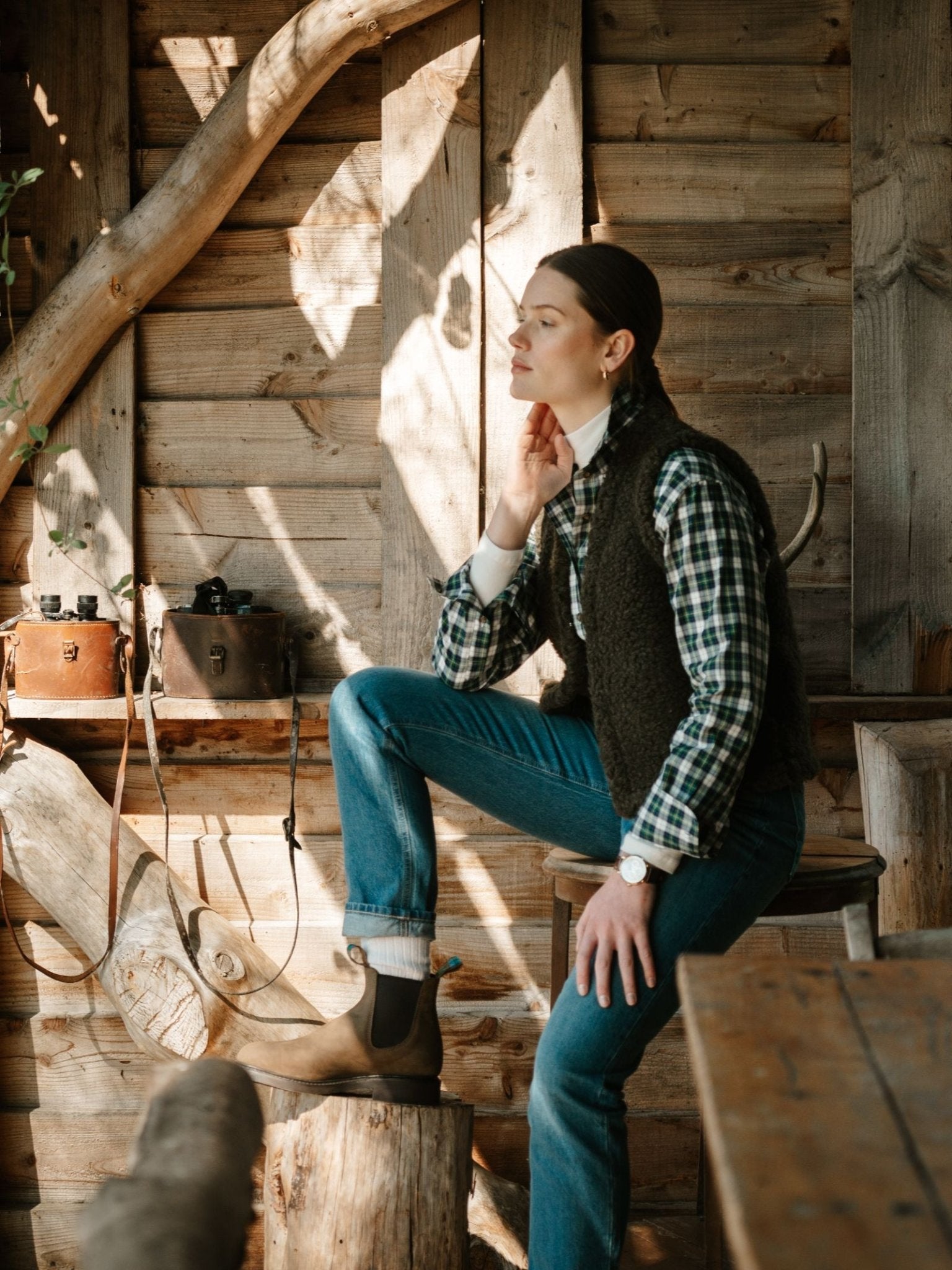 A woman in a plaid shirt, vest, and jeans sits indoors on a wooden stool, her foot in the R.M.Williams x Campbell's of Beauly Ladies' Highlander Boot resting on a log. Vintage cameras and wood walls surround her as she gazes thoughtfully.