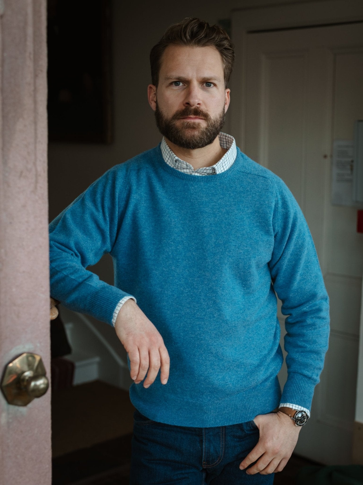 A bearded man wearing a Campbell's of Beauly Lambswool Crew Neck Jumper in blue over a collared shirt and jeans stands indoors by a door, one hand on the frame, looking directly at the camera.