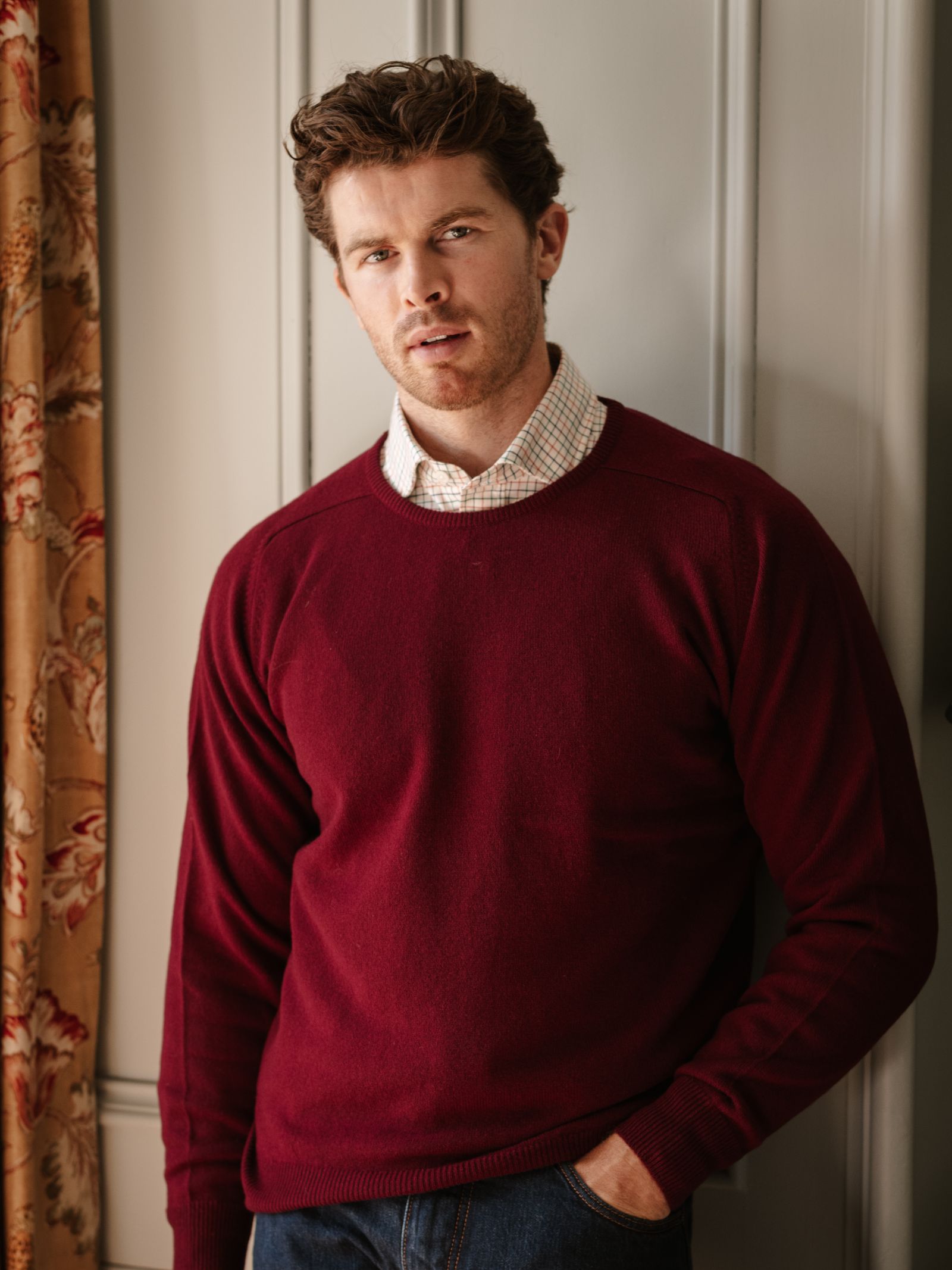 A man with short, curly brown hair and a beard wears a Campbell's of Beauly Lambswool Crew Neck Jumper over a plaid shirt, standing indoors with one hand in his pocket by a paneled wall and floral curtain.
