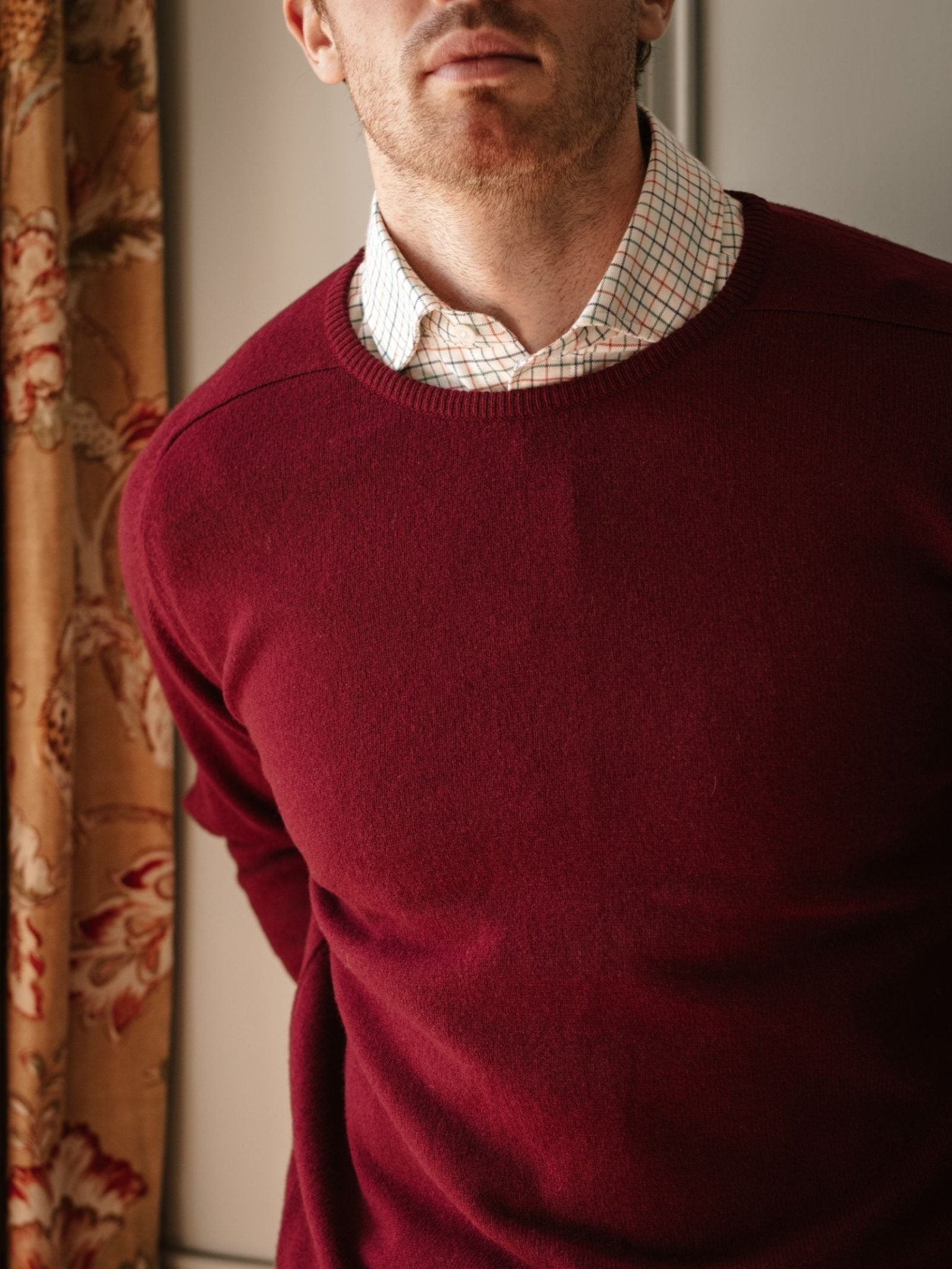 A man, visible from torso to lower face, stands indoors with arms behind his back, wearing a Campbell's of Beauly Lambswool Crew Neck Jumper in red over a white checkered shirt near a floral curtain.