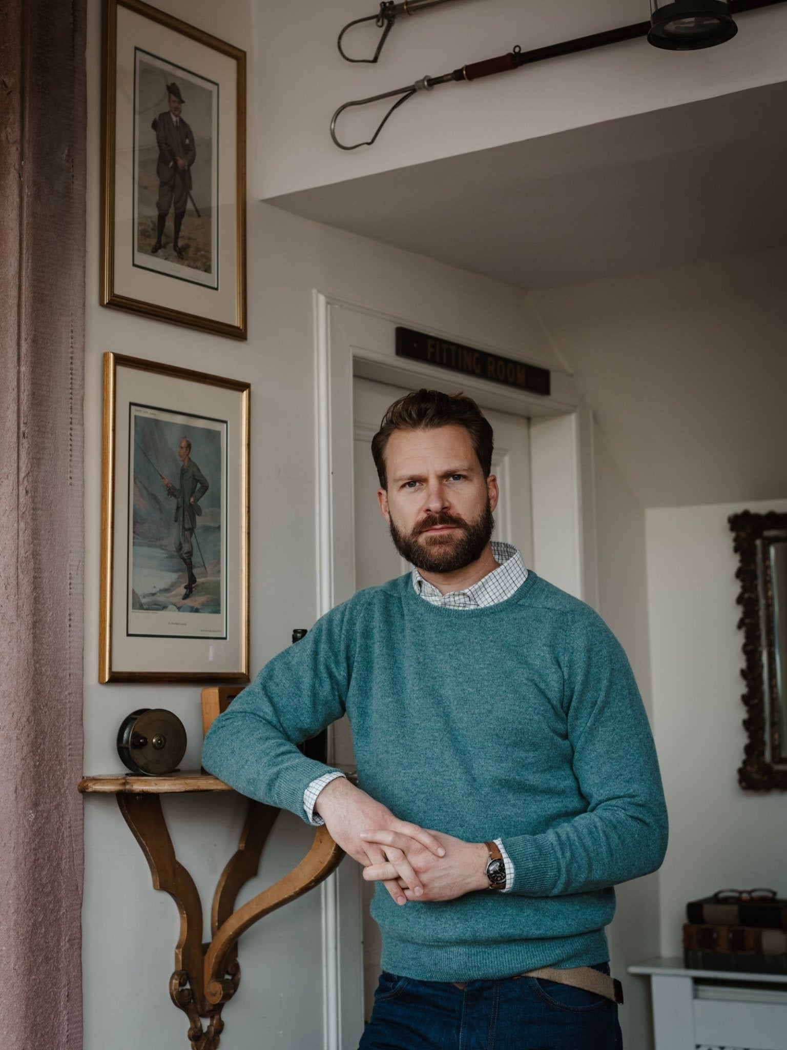 A bearded man wears the Lambswool Crew Neck Jumper by Campbell's of Beauly over a collared shirt, standing indoors beside a small shelf, framed art, vintage decor, and a Fitting Room sign in the background.